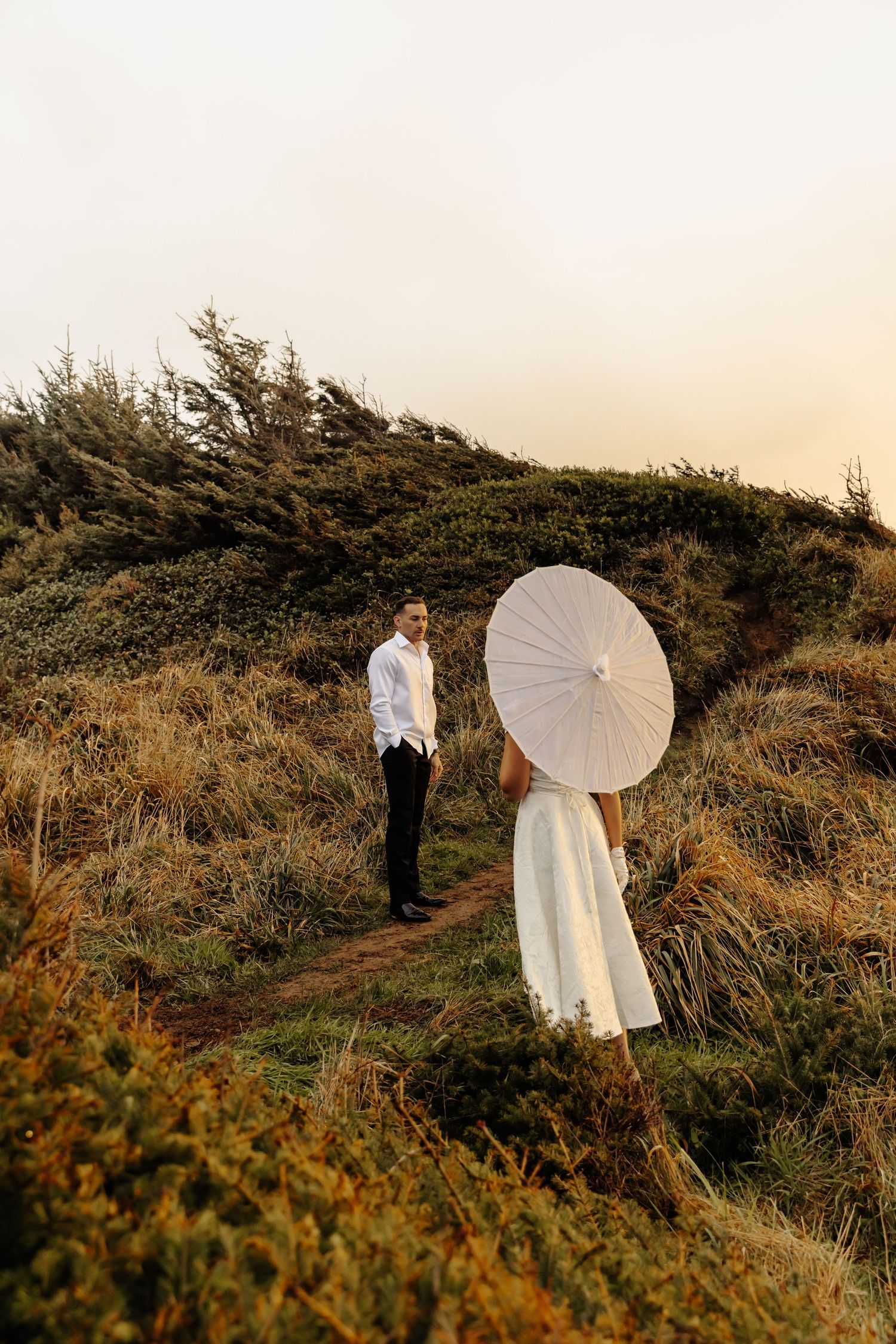 A couple poses with a white parasol on a grassy hillside overlooking the ocean.