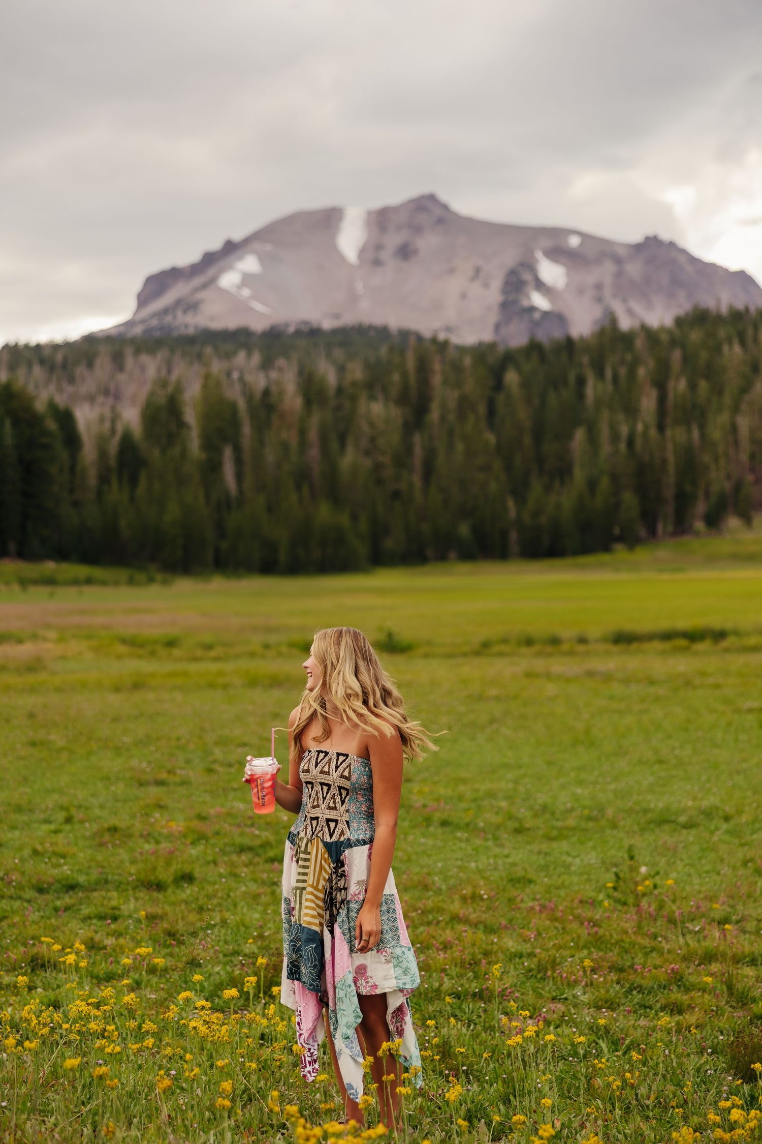 Blonde woman in colorful boho dress holding drink stands in wildflower meadow with pine forest and snowy mountain backdrop.