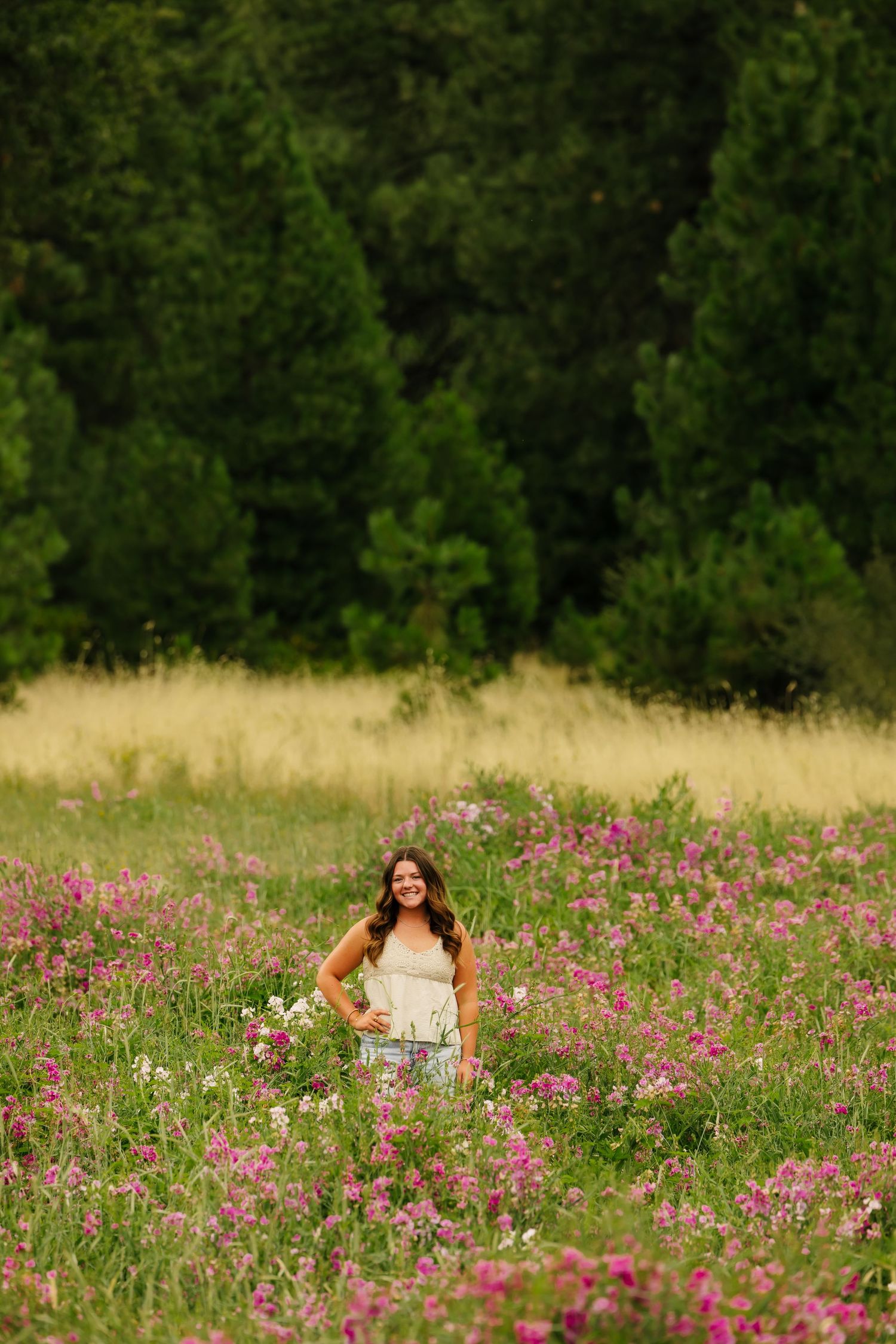 Young woman in white dress smiling in a vibrant wildflower meadow with pink blooms and lush green forest backdrop.