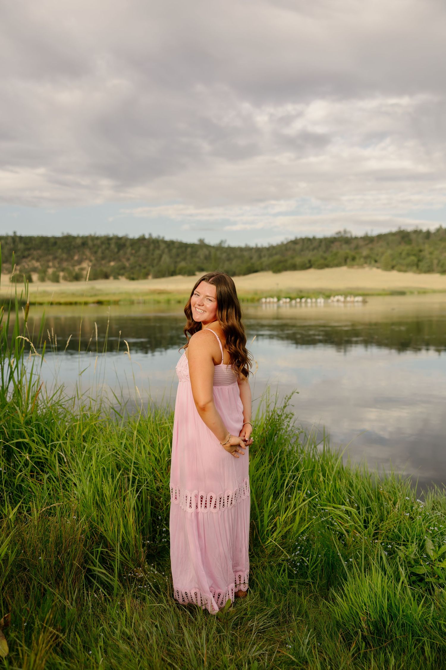 Young woman in pink maxi dress smiling by a reflective lake with lush green grass and cloudy sky.