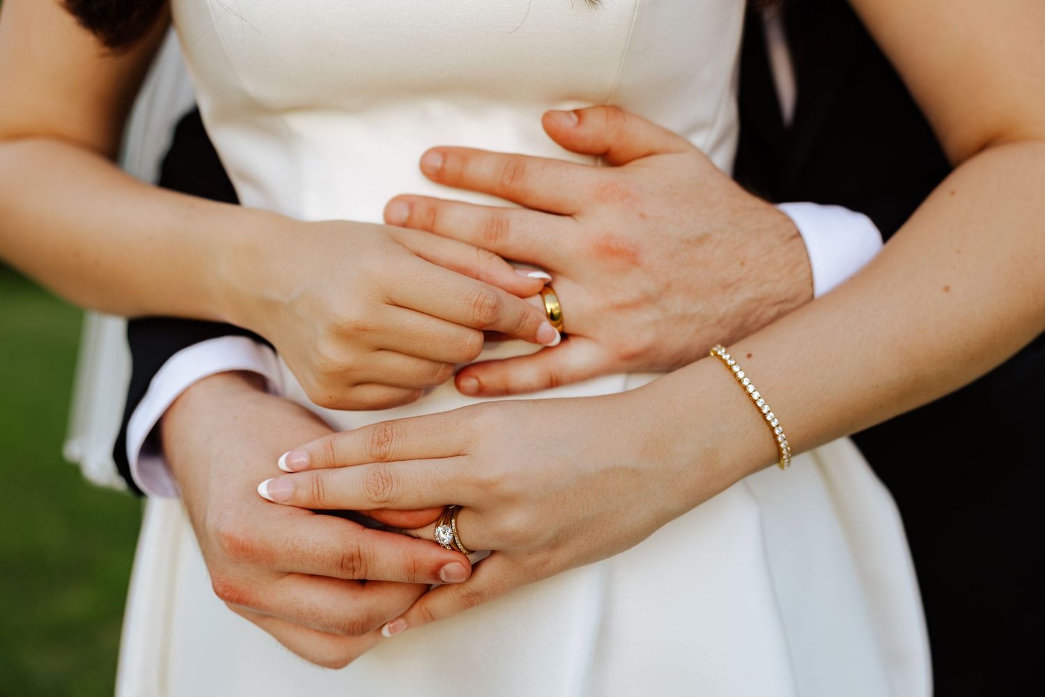Close-up of newlywed couple hands showing wedding rings, bride wearing gold bracelet and white dress.