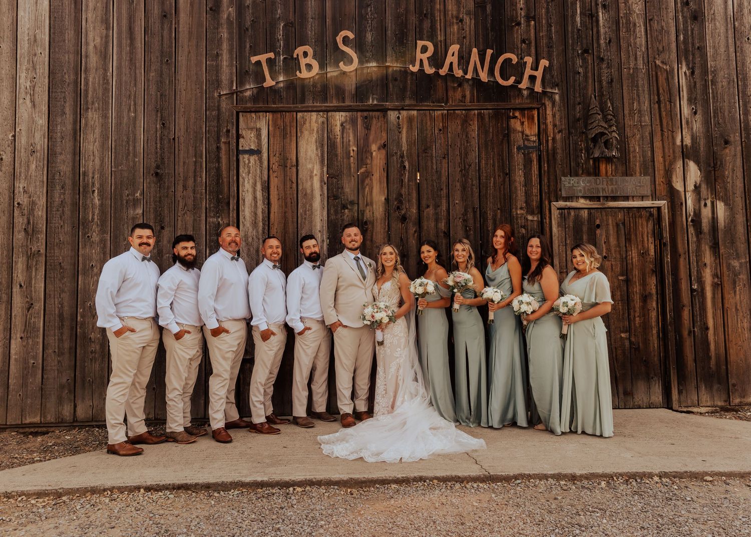 Wedding party group photo in front of rustic wooden barn with TRS Ranch signage.