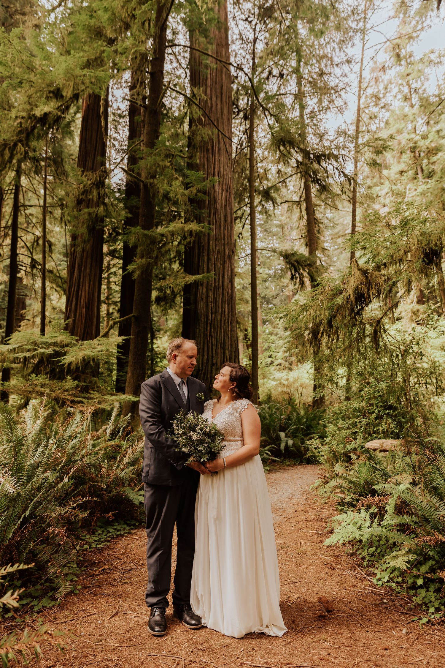 A couple shares an intimate moment on a dirt path through a majestic redwood forest.