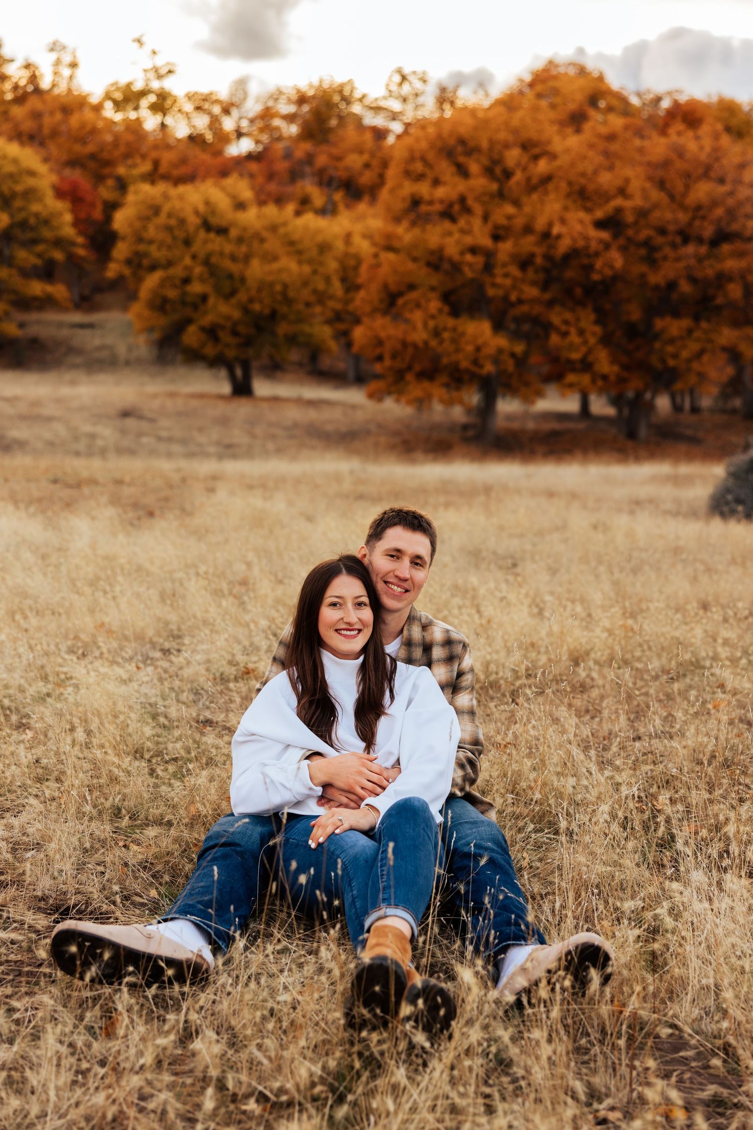 Couple sitting together in a golden field with autumn trees behind them.