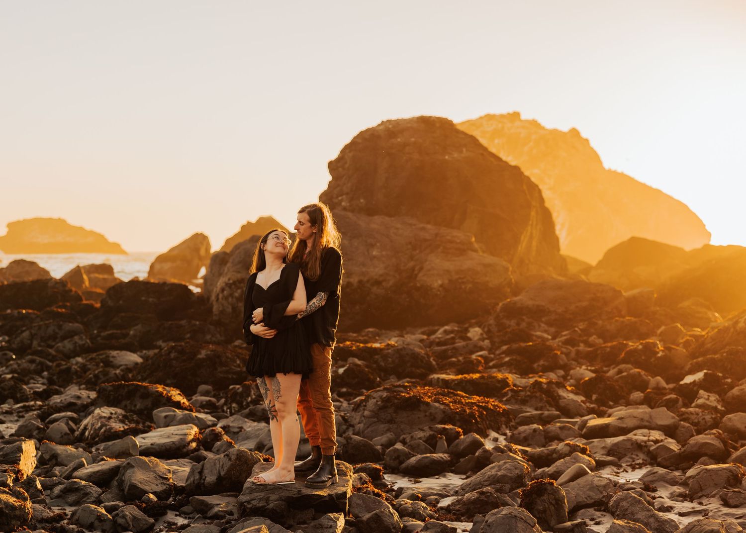 Couple embraces on rocky beach during golden sunset with dramatic coastal rock formations in background.