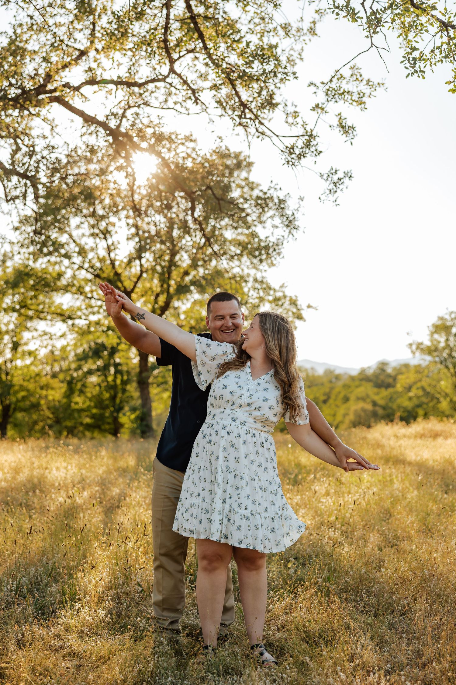 Couple dancing in golden field at sunset under oak tree, woman in floral dress, man in navy shirt.