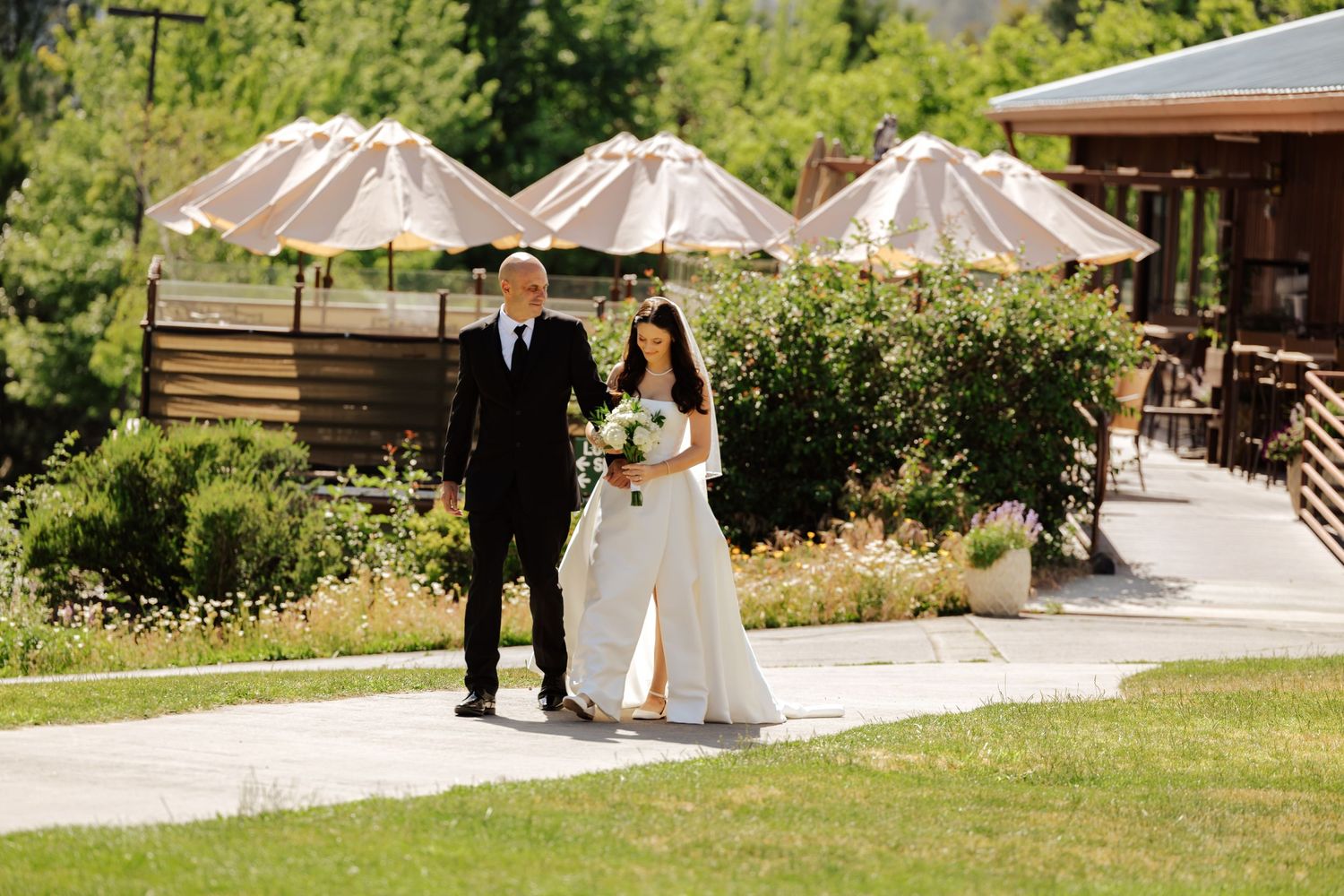 Bride in white gown walks with escort on sunny path at outdoor wedding venue with umbrellas and greenery.
