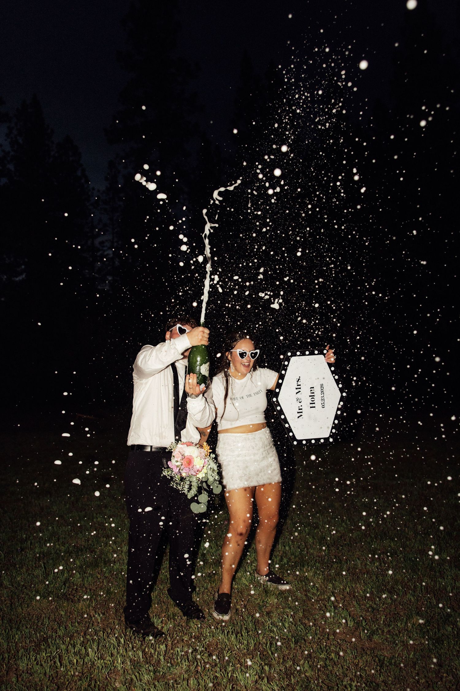 Newlyweds celebrate at night popping champagne bottle with confetti flying, bride holding bouquet in white dress.