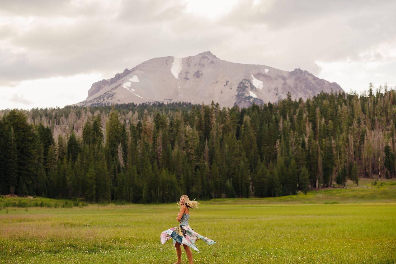 Woman in flowing dress walking in green meadow with dense pine forest and snow-capped mountain peak backdrop.