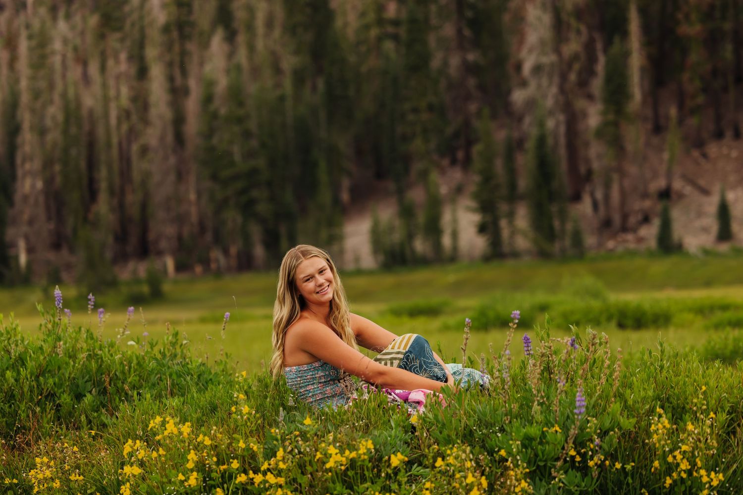 Young blonde woman smiling in a wildflower meadow with tall pine trees in the background.
