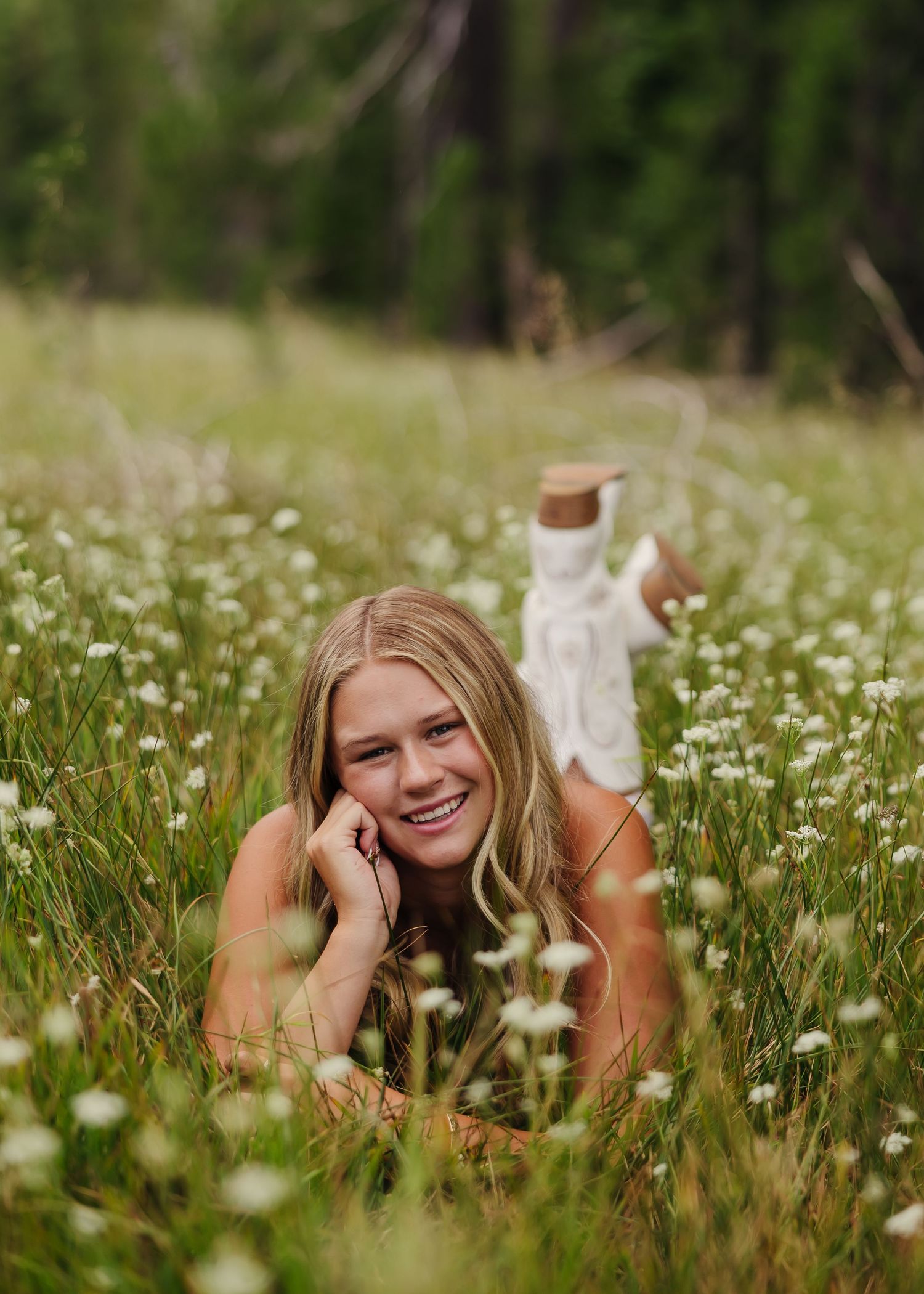 Smiling blonde woman lying in a wildflower meadow surrounded by white daisies and lush green grass.