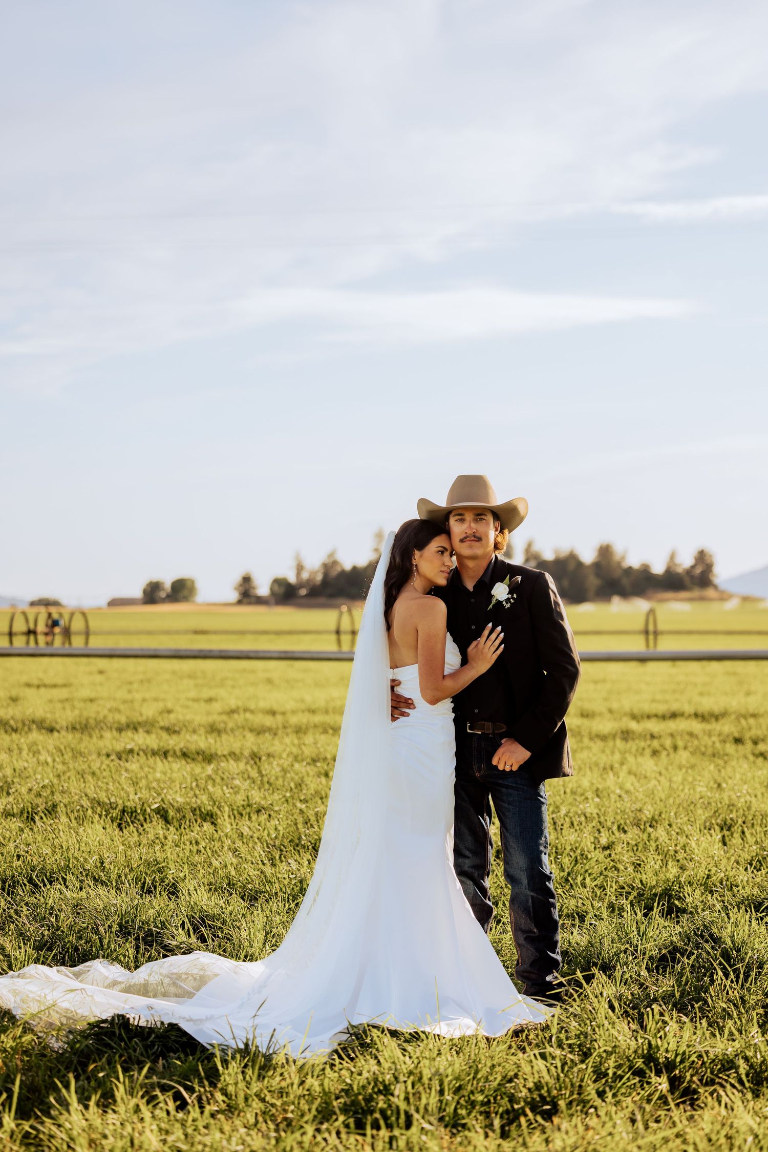 Bride in white gown and groom in cowboy hat embrace in a lush green field at golden hour.