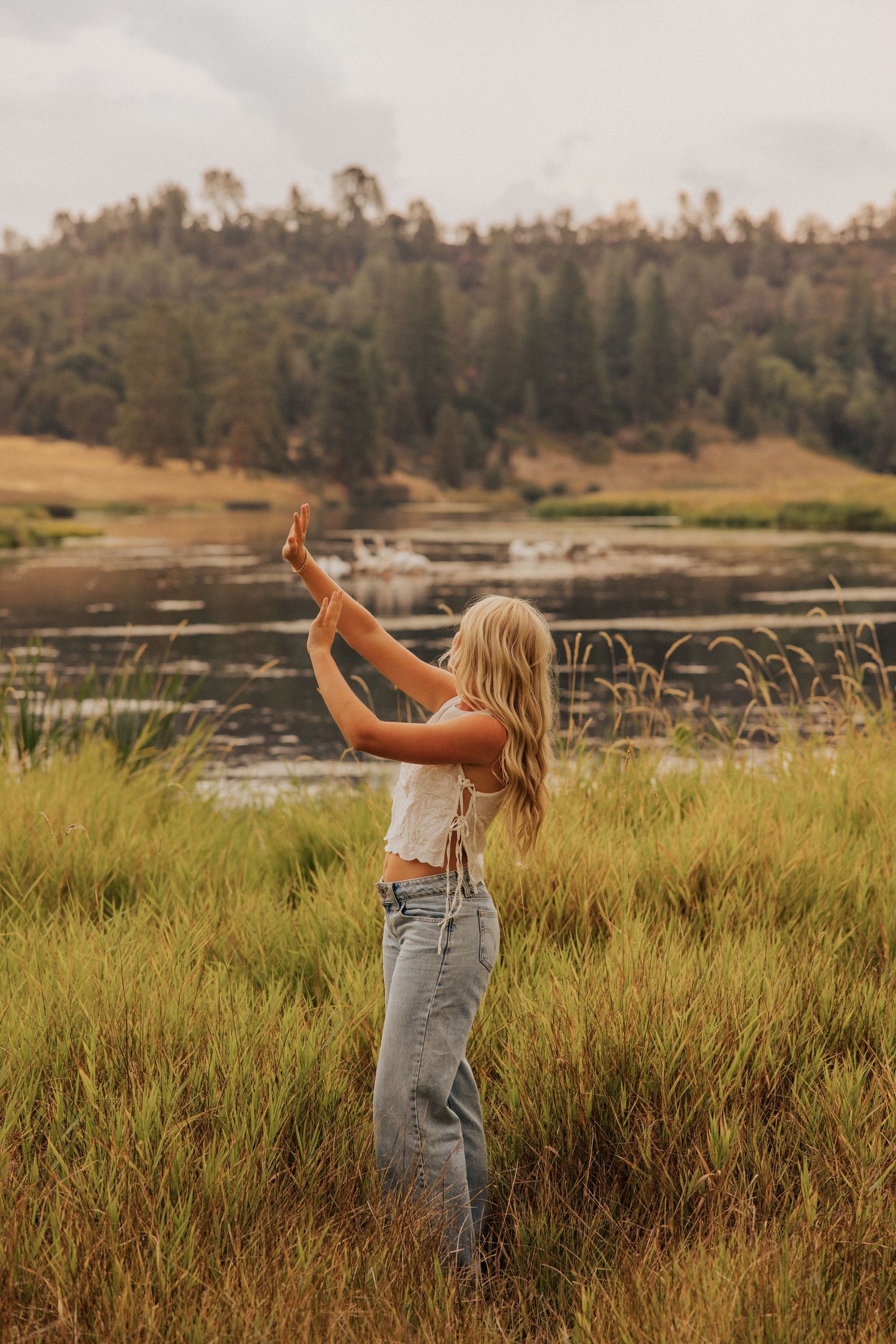 Person in casual outfit dancing in tall grass with lake and mountains in background.