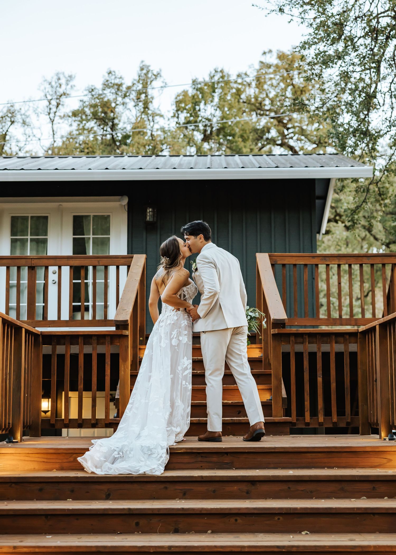 Bride and groom share a kiss on a wooden deck outside a dark cabin venue during their outdoor wedding celebration.