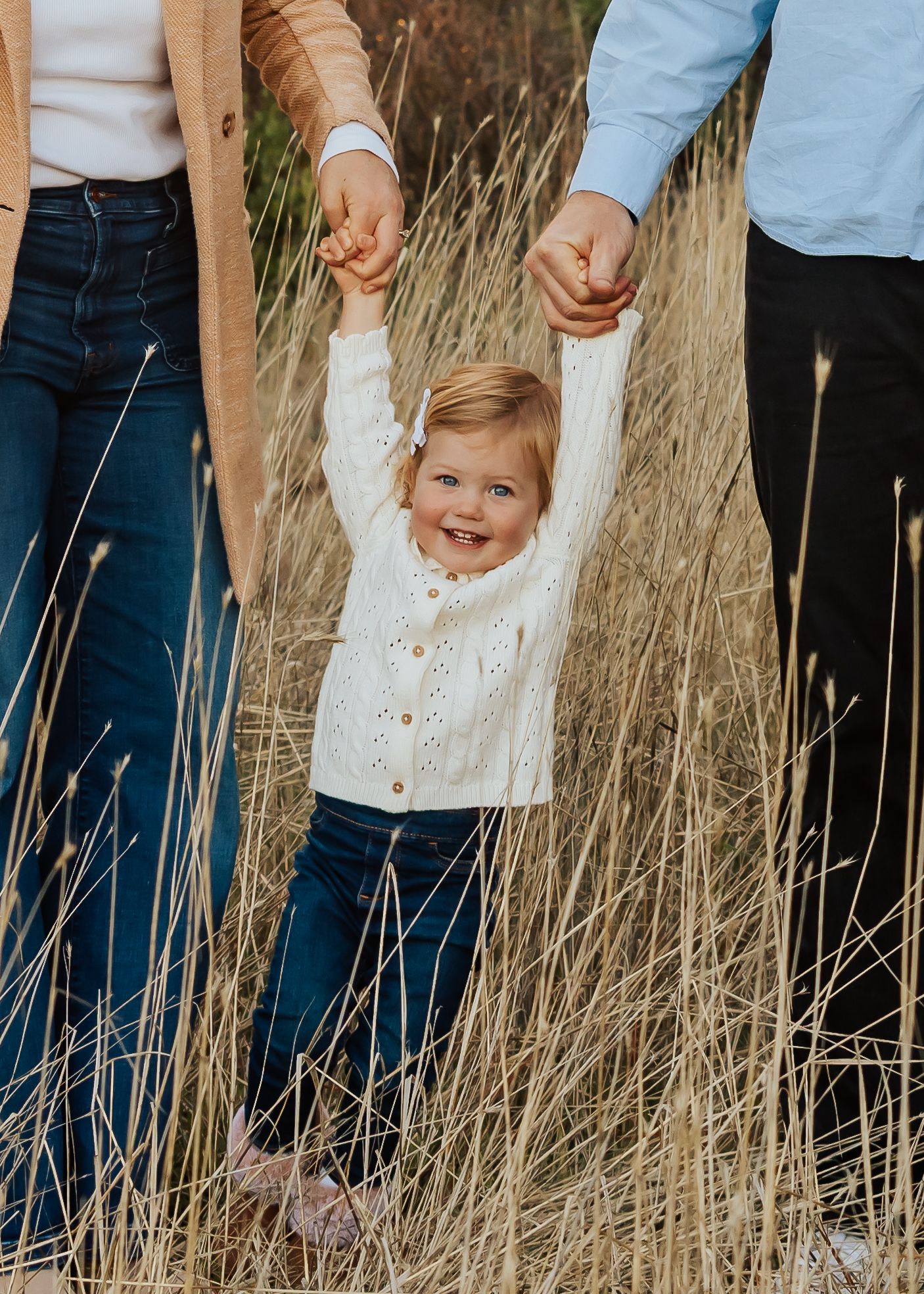 Happy toddler swings between parents' hands in tall grass field.