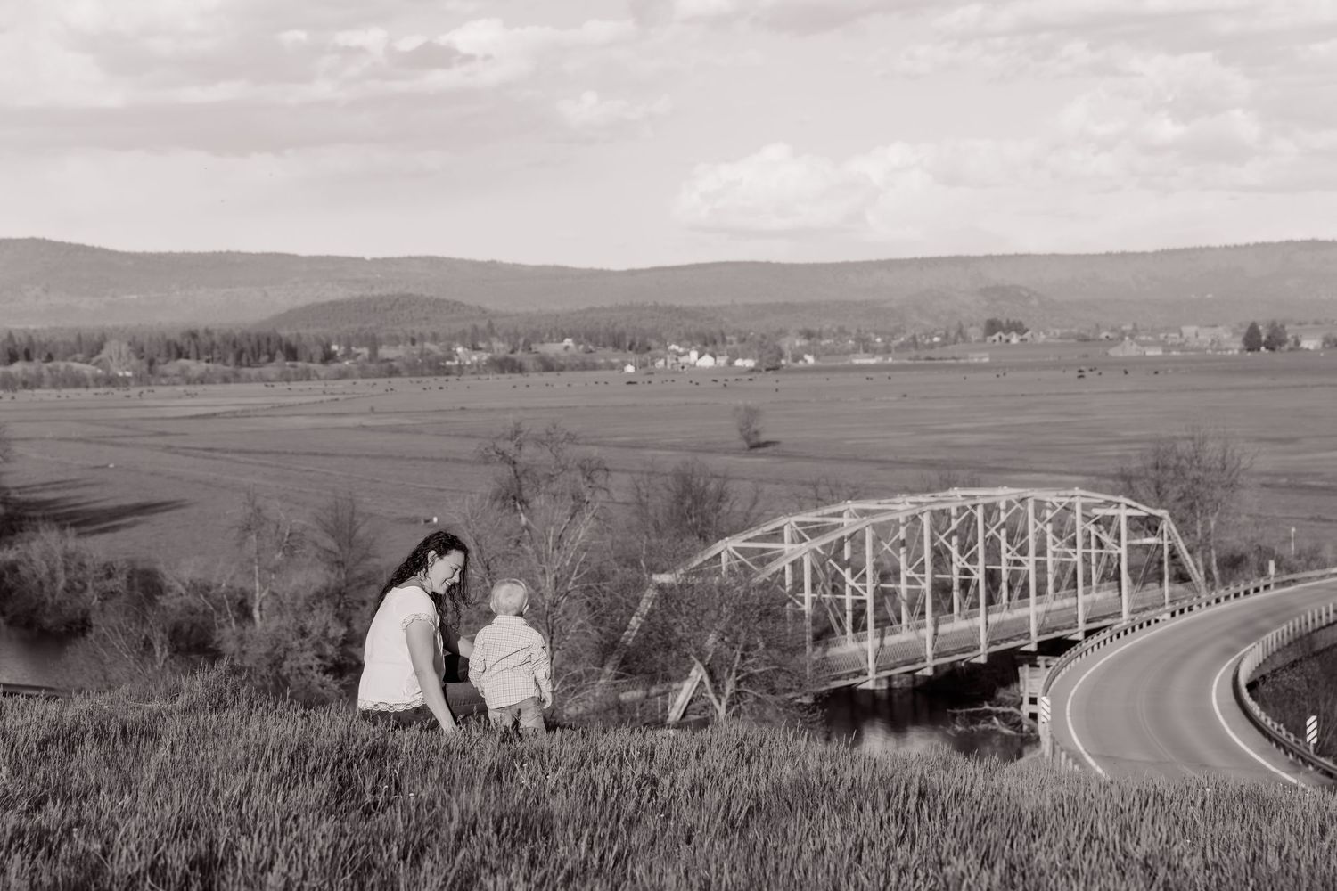 Rural landscape with winding road, bridge, and rolling hills in black and white.