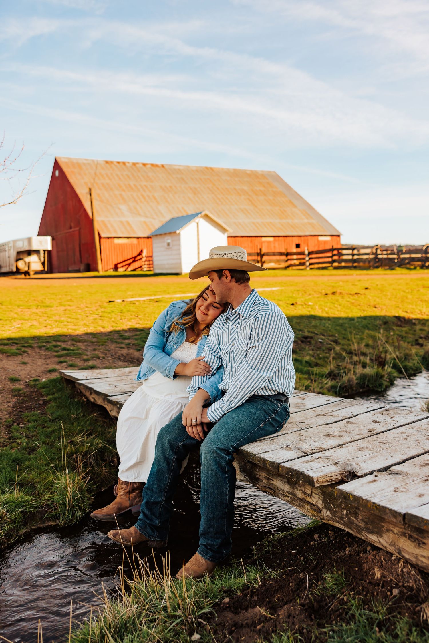 A romantic couple sits together on wooden bridge with red barn in background.