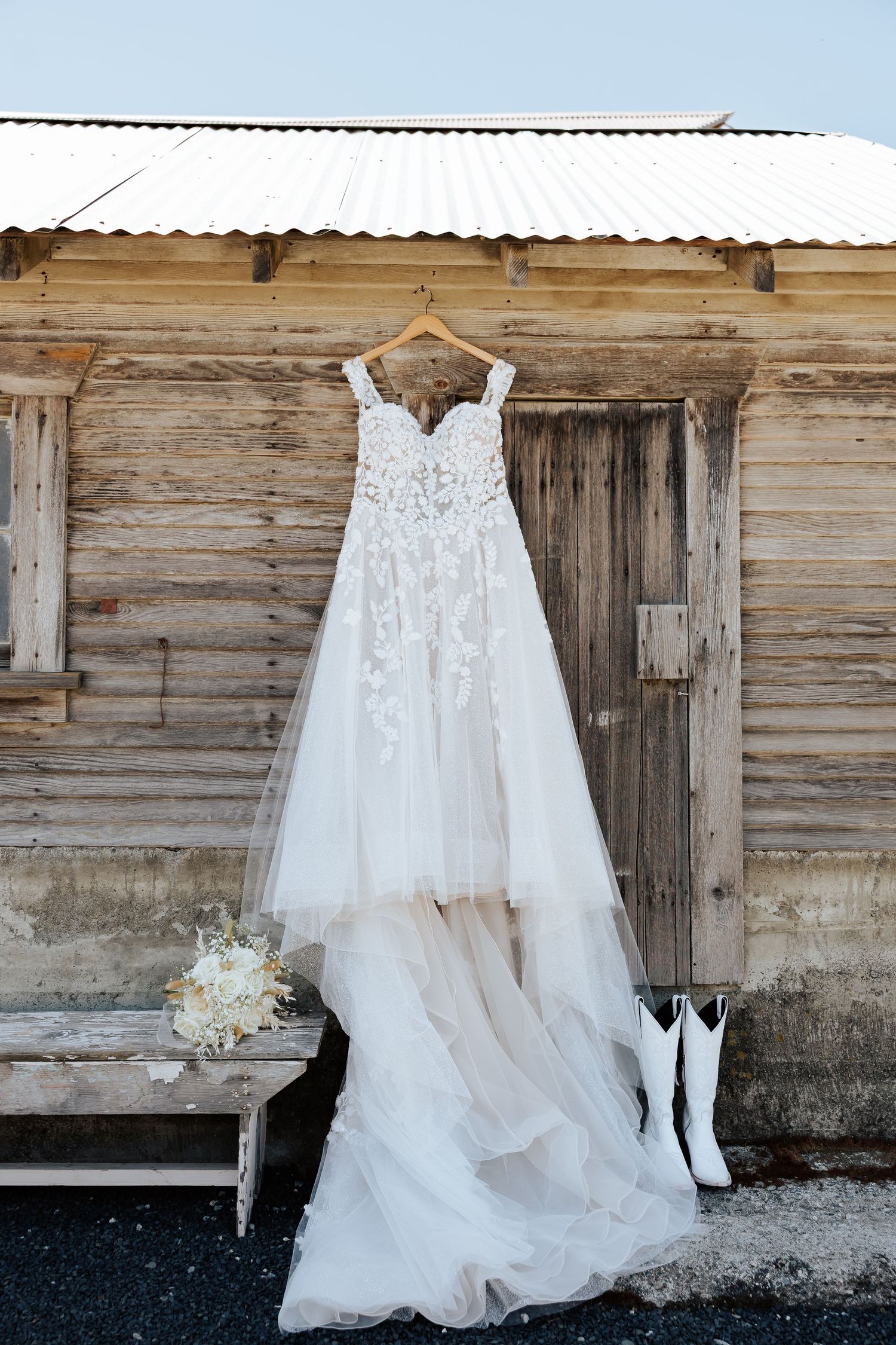 A lace wedding dress hangs outside a rustic wooden barn with white cowboy boots and a floral bouquet below.