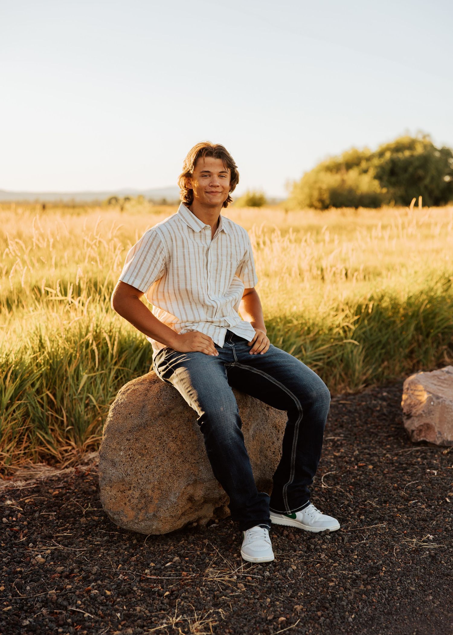 A person sits on a large rock at sunset wearing jeans and a casual shirt.