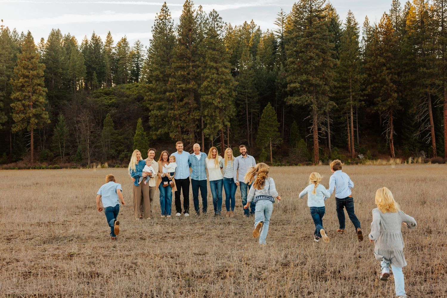 Candid family photo showing group running and playing in a rustic field with evergreen forest.