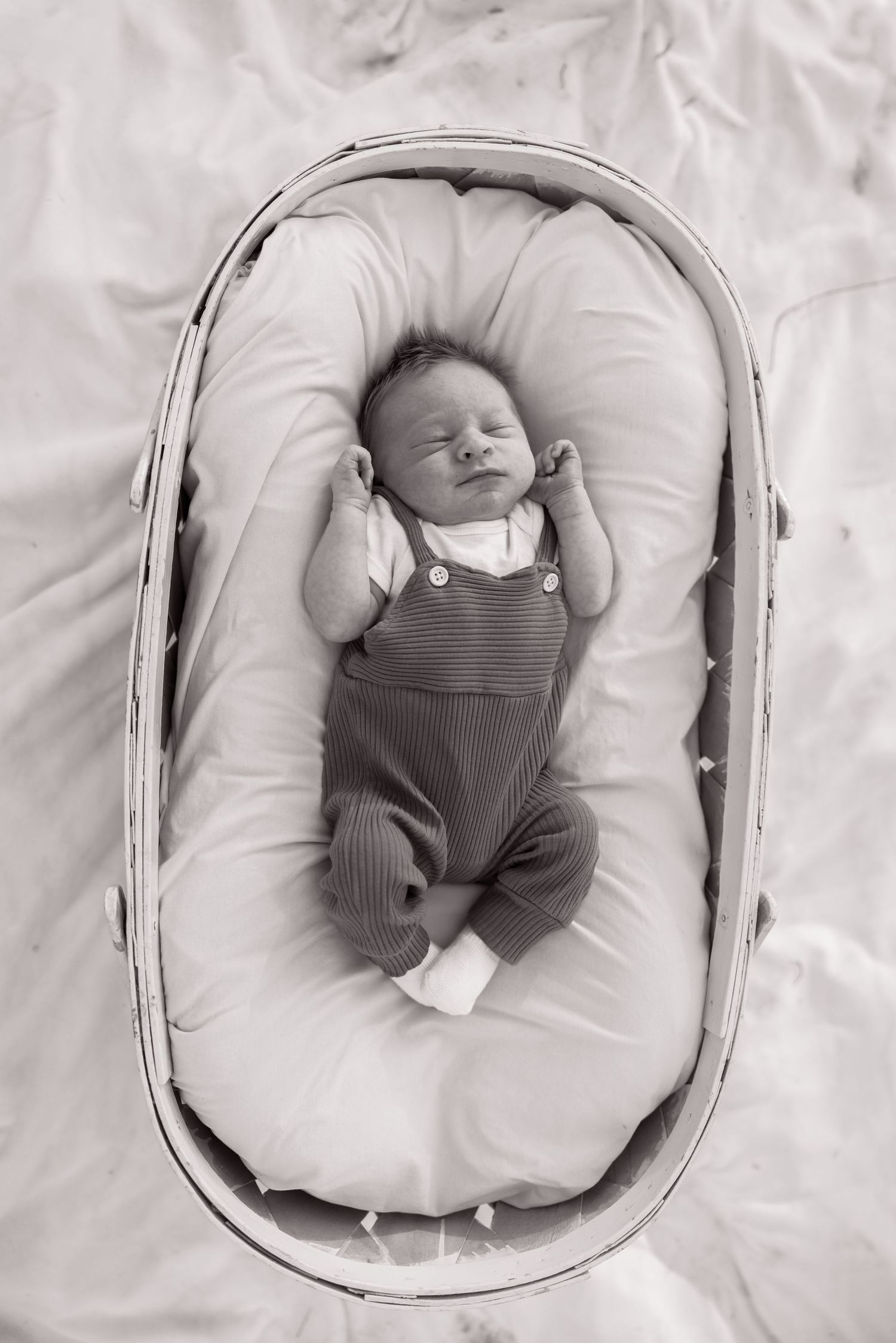 A sleeping newborn baby wearing overalls rests peacefully in a white cushioned bassinet.