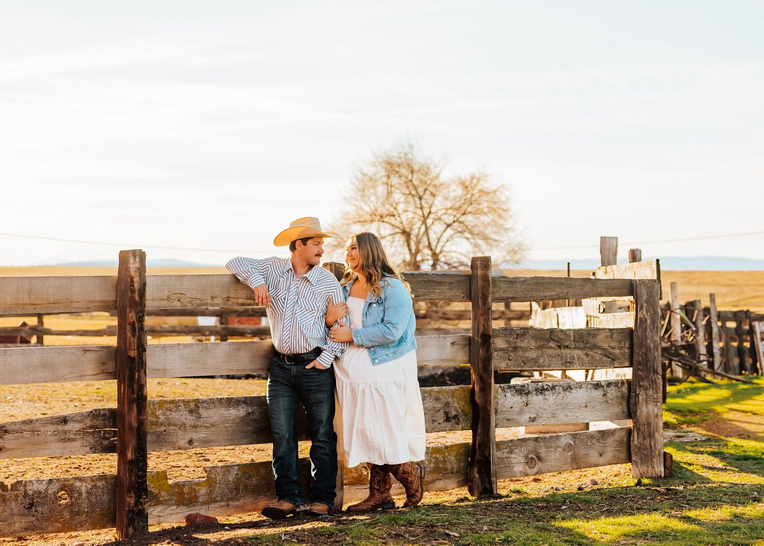 A couple in western attire poses together by a rustic wooden fence at sunset on a ranch.