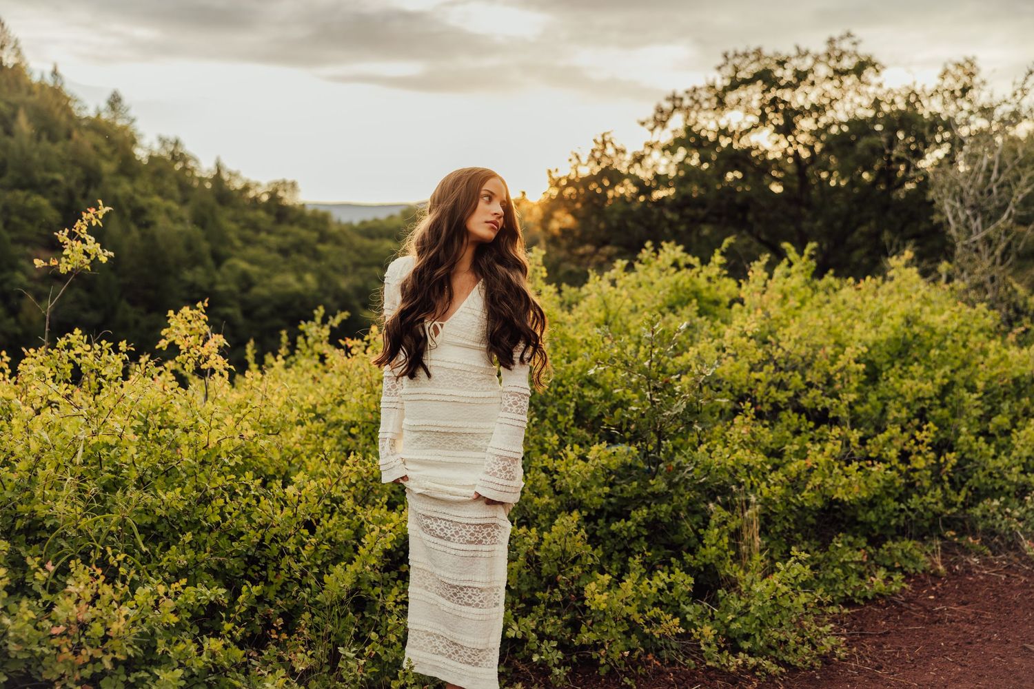 A person in a flowing white dress stands among yellow flowers with mountains in the distance.