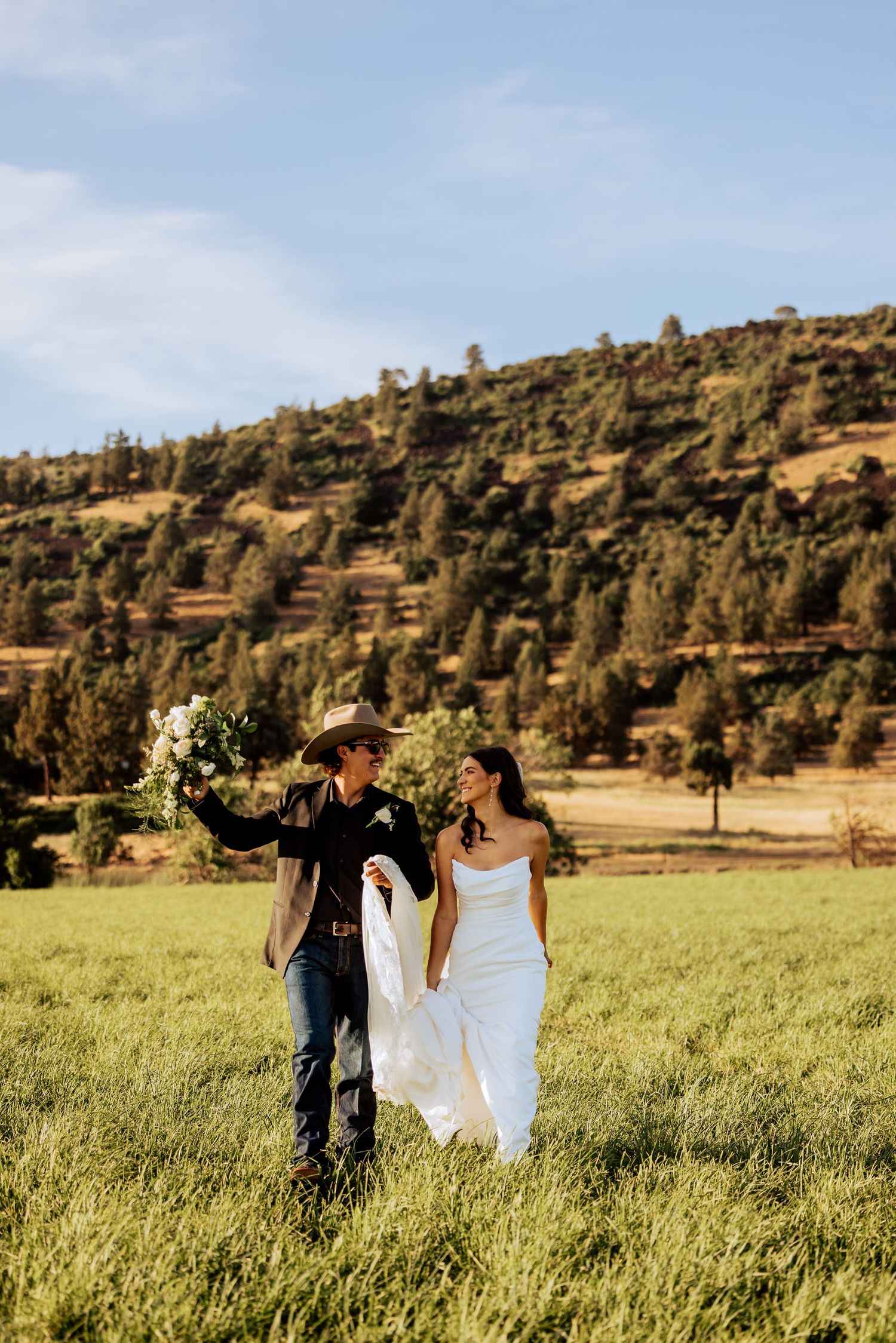 Newlyweds walk hand in hand through a scenic meadow with hills in the background.