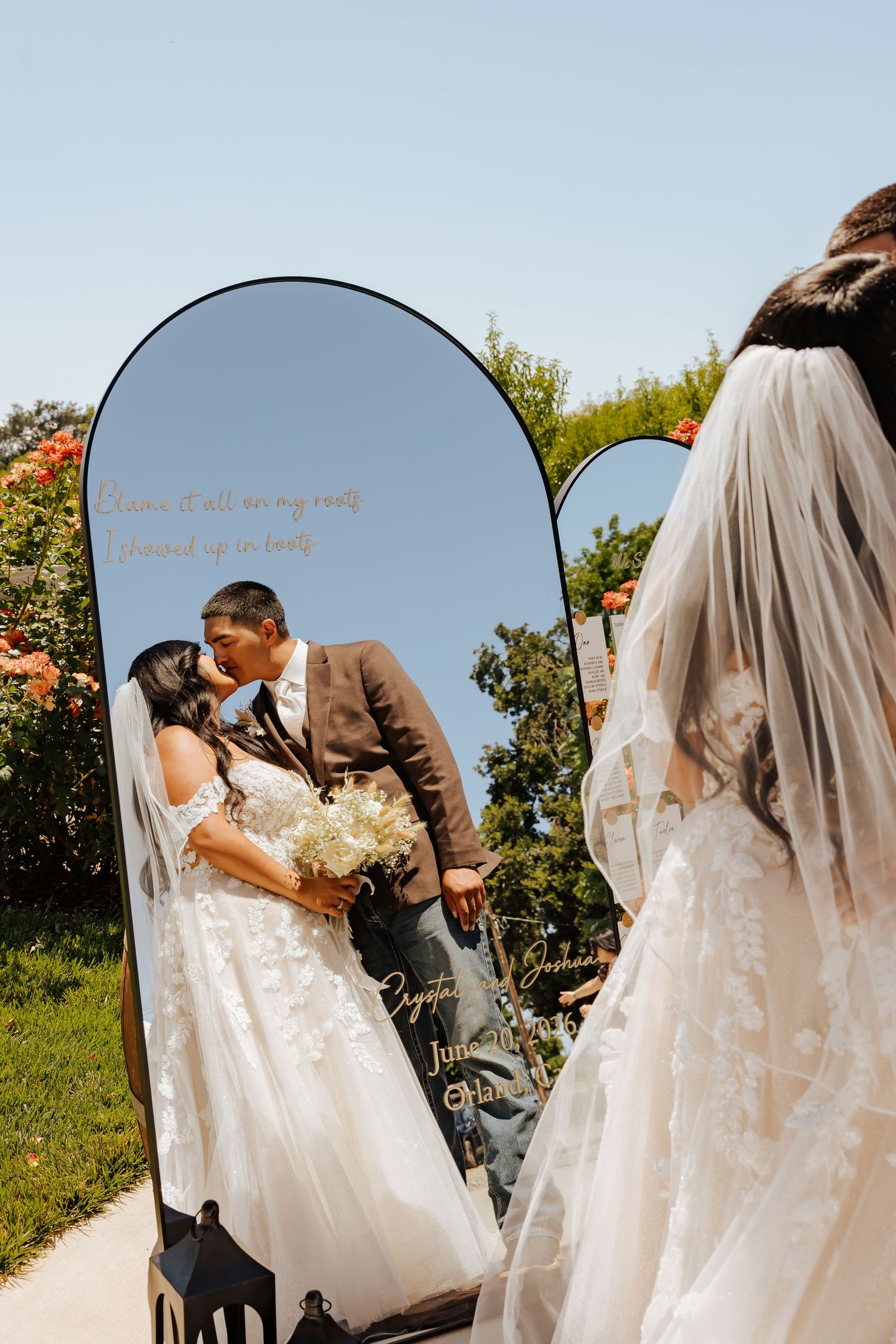 Bride and groom share a kiss reflected in an elegant arched mirror at an outdoor wedding ceremony.