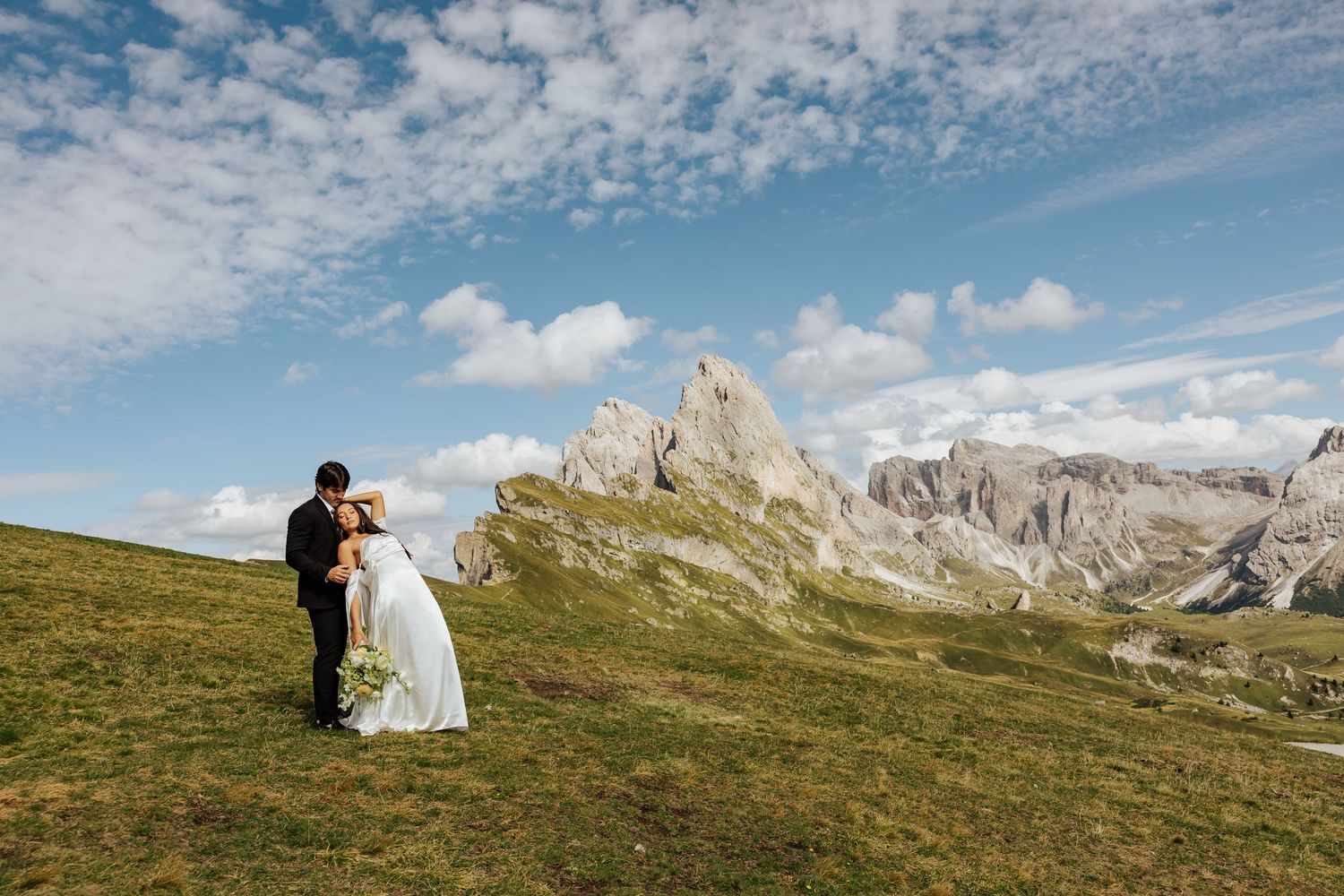 Bride and groom embrace on a lush green hillside with dramatic Dolomite mountain peaks under a blue sky.