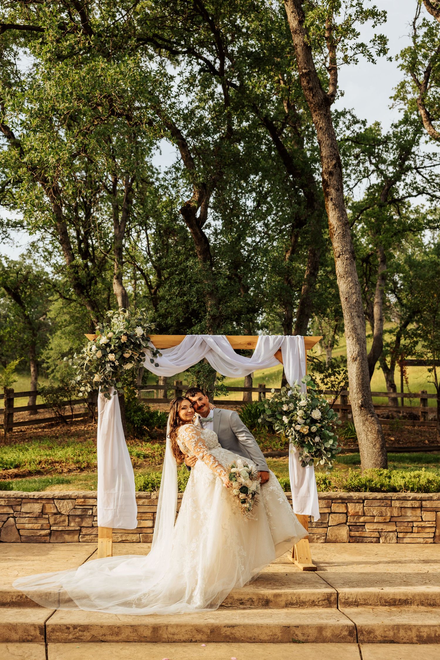 Bride and groom pose under a white draped floral arch at an elegant outdoor wedding ceremony surrounded by lush trees.