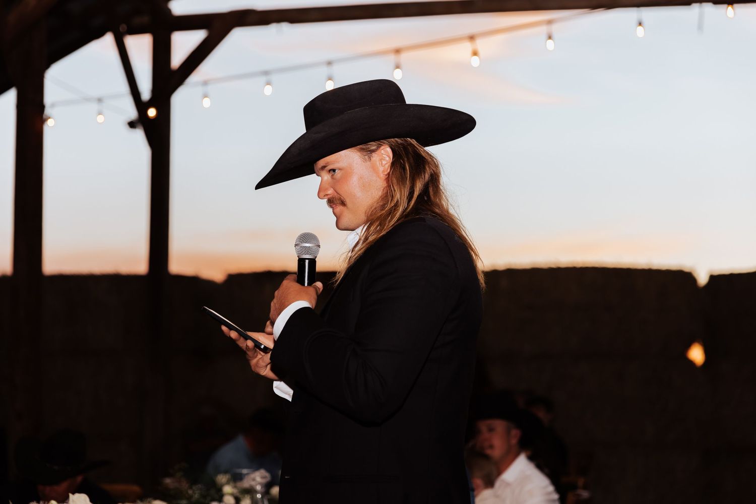 Person in black suit and cowboy hat speaking into microphone at outdoor wedding reception at dusk.