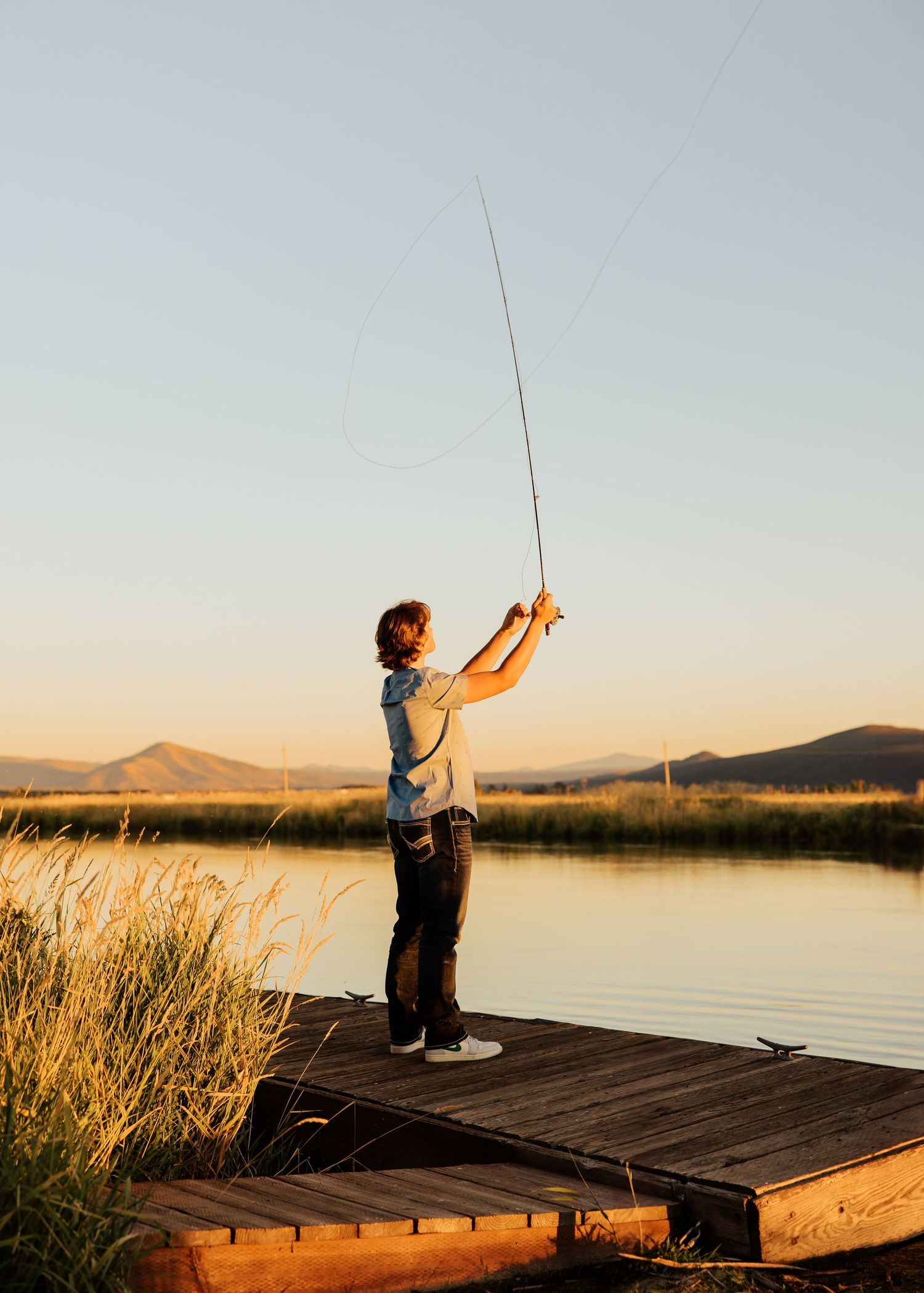 A silhouette of a person fishing on a wooden dock at sunset with mountains in the background.