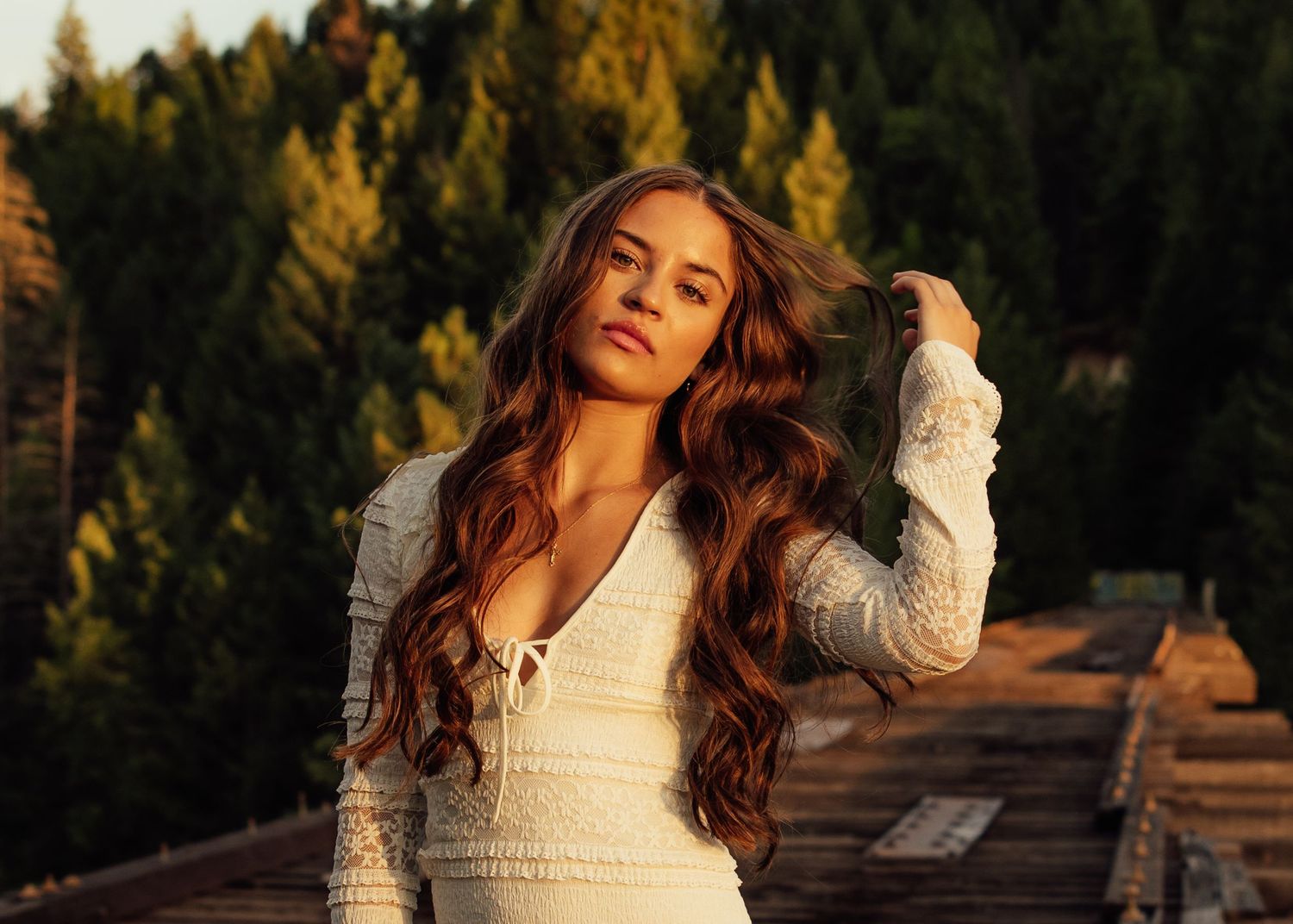 A person in a white lace dress stands against a backdrop of evergreen trees at sunset.