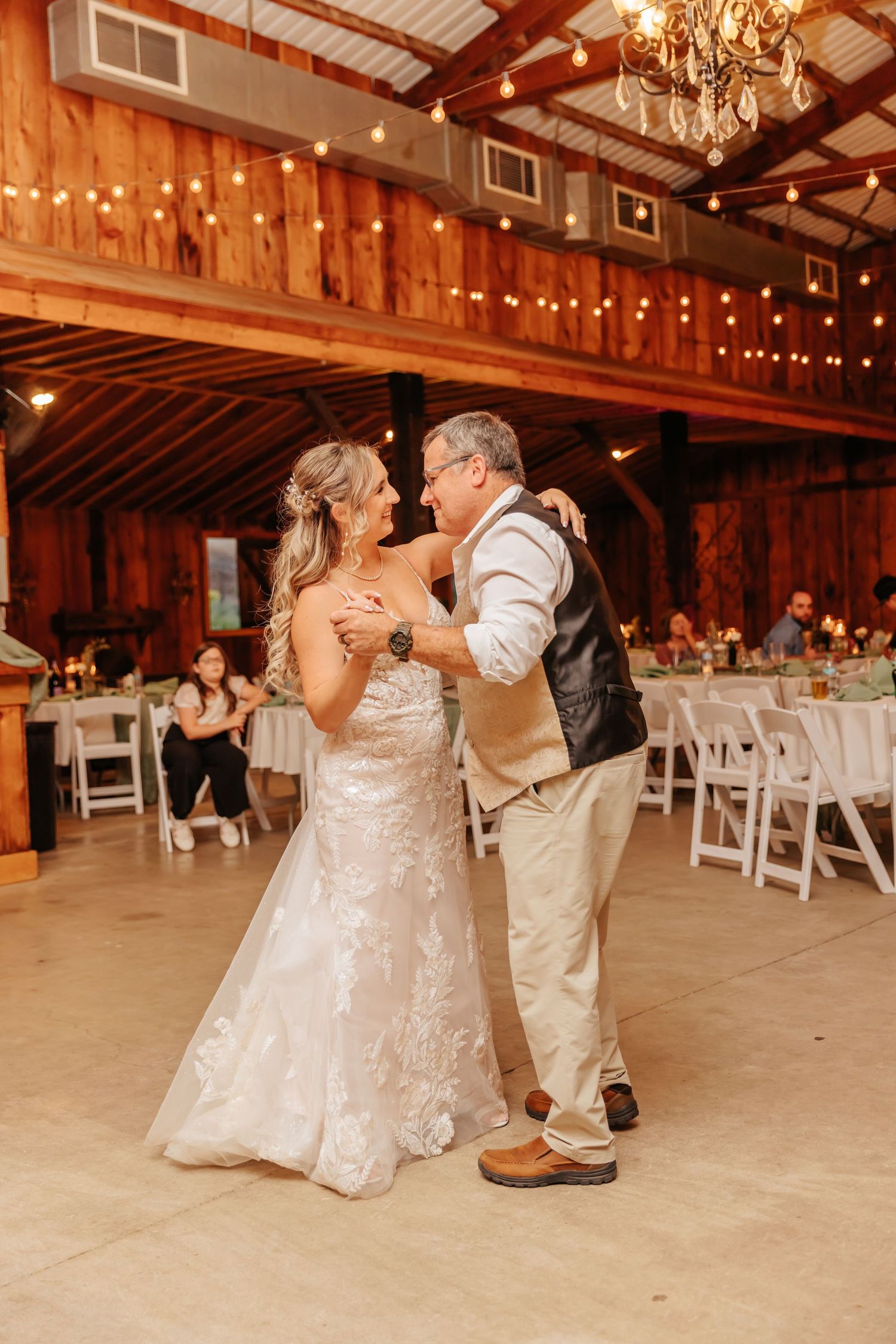 Wedding couple dances together in beautifully lit barn venue with white chairs and string lights.