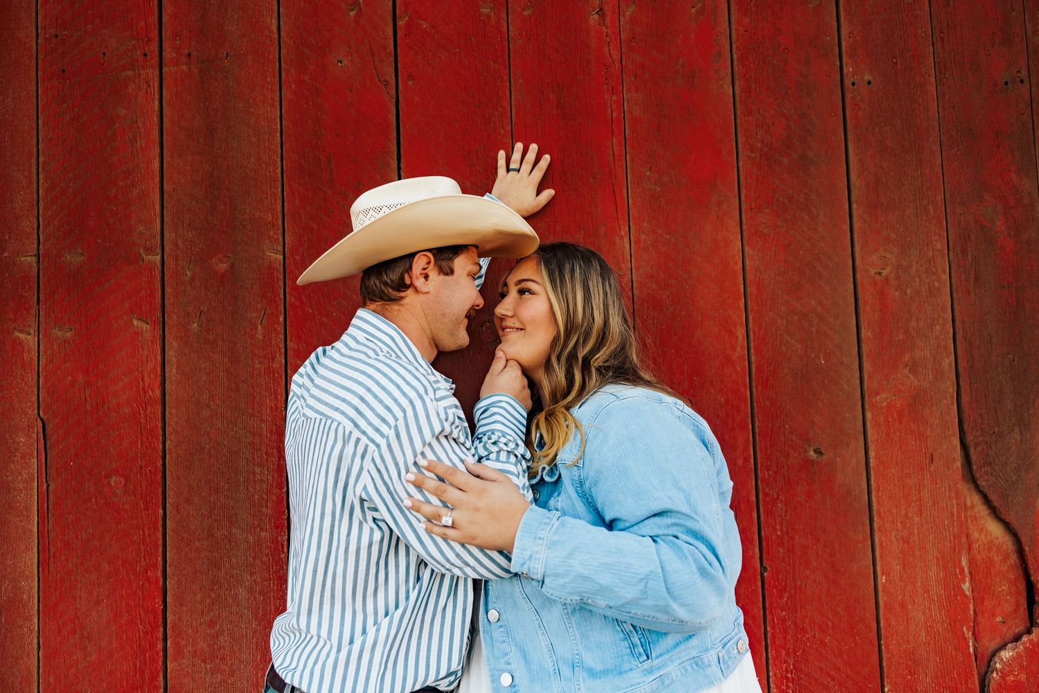 A romantic couple embraces against a rustic red barn wall wearing casual western attire.