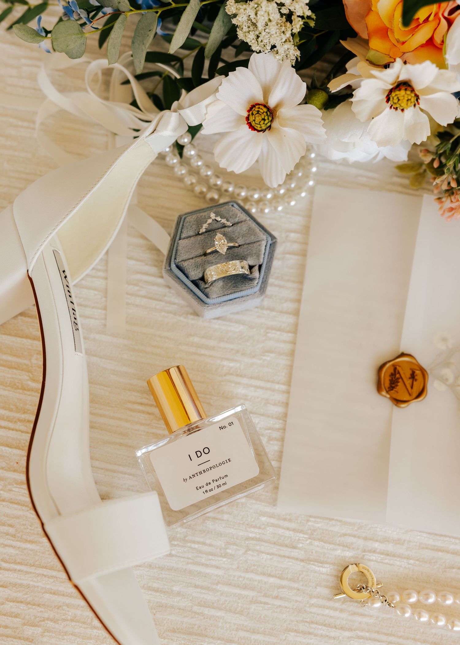 Wedding details including white flowers, pearls and perfume bottles arranged on textured fabric.