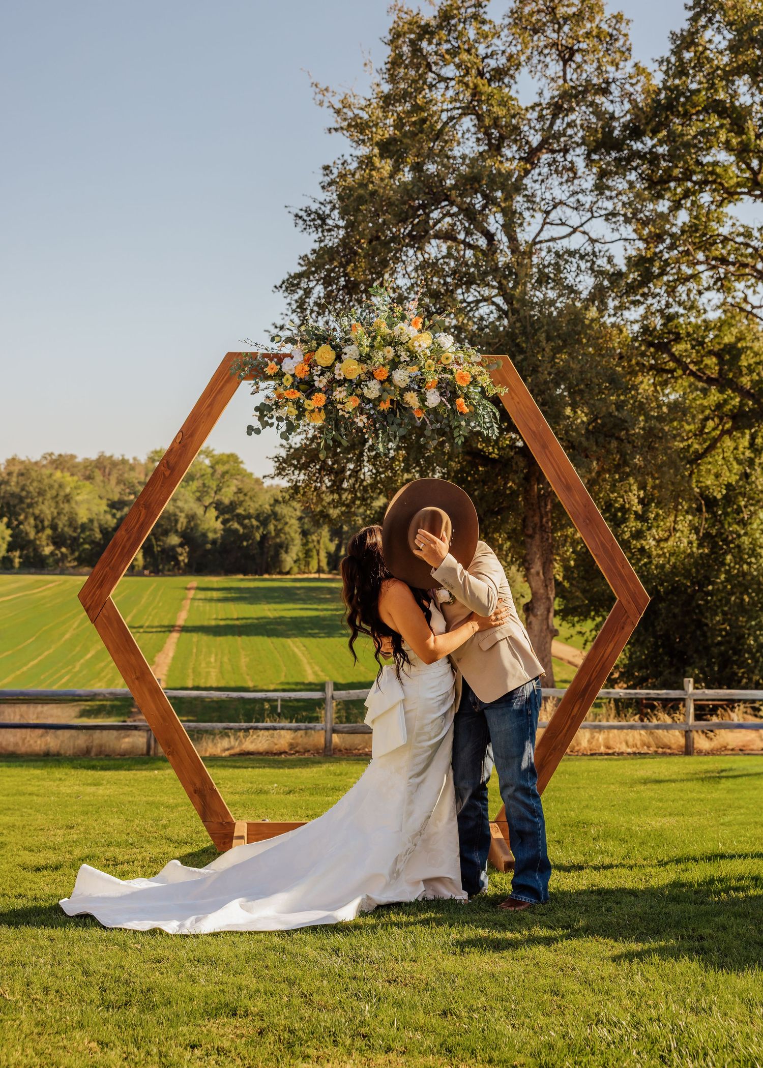 Wedding couple poses by wooden hexagon arch in sunny outdoor field setting.