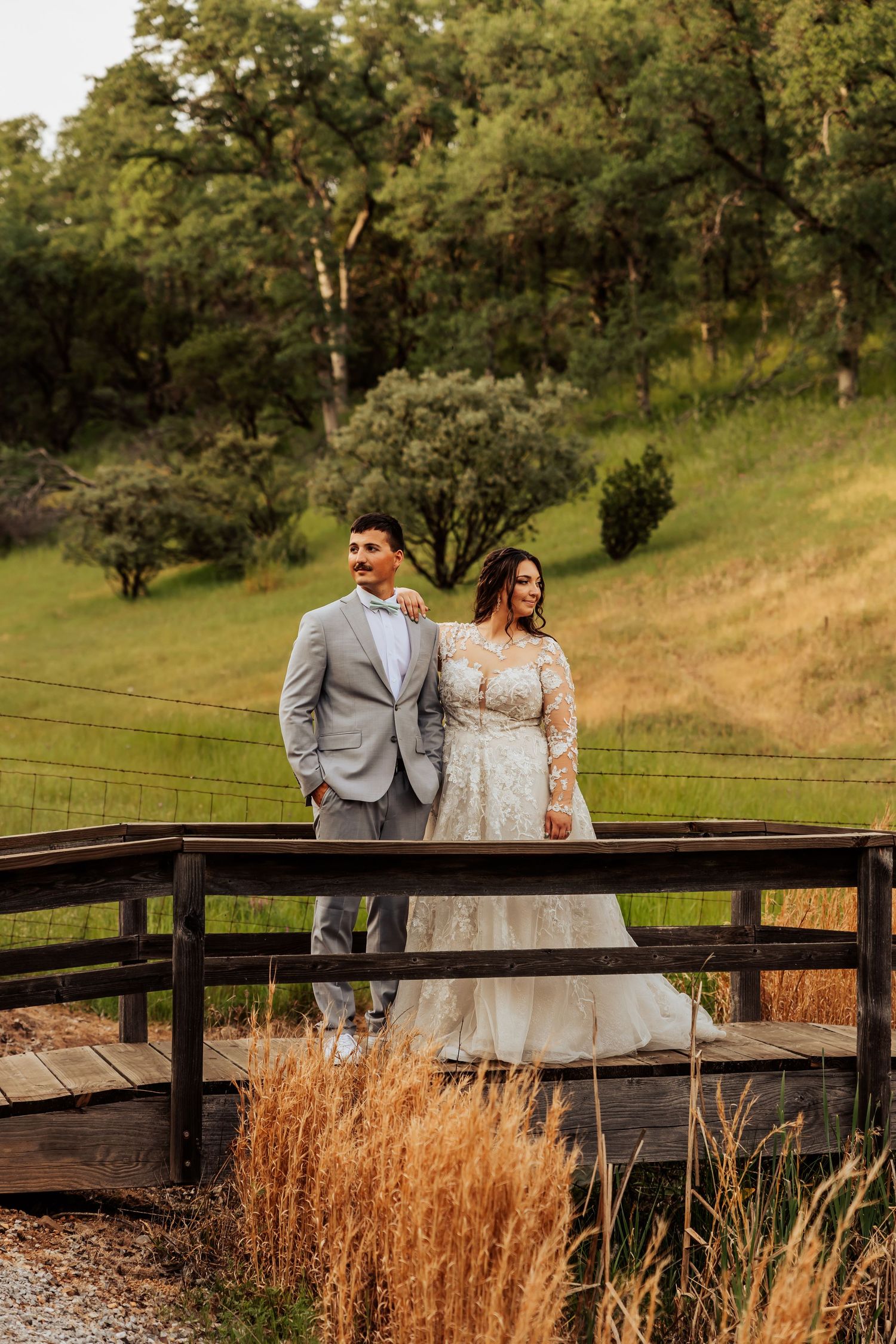 Bride and groom standing on a wooden bridge surrounded by lush green hills and golden grasses during golden hour.