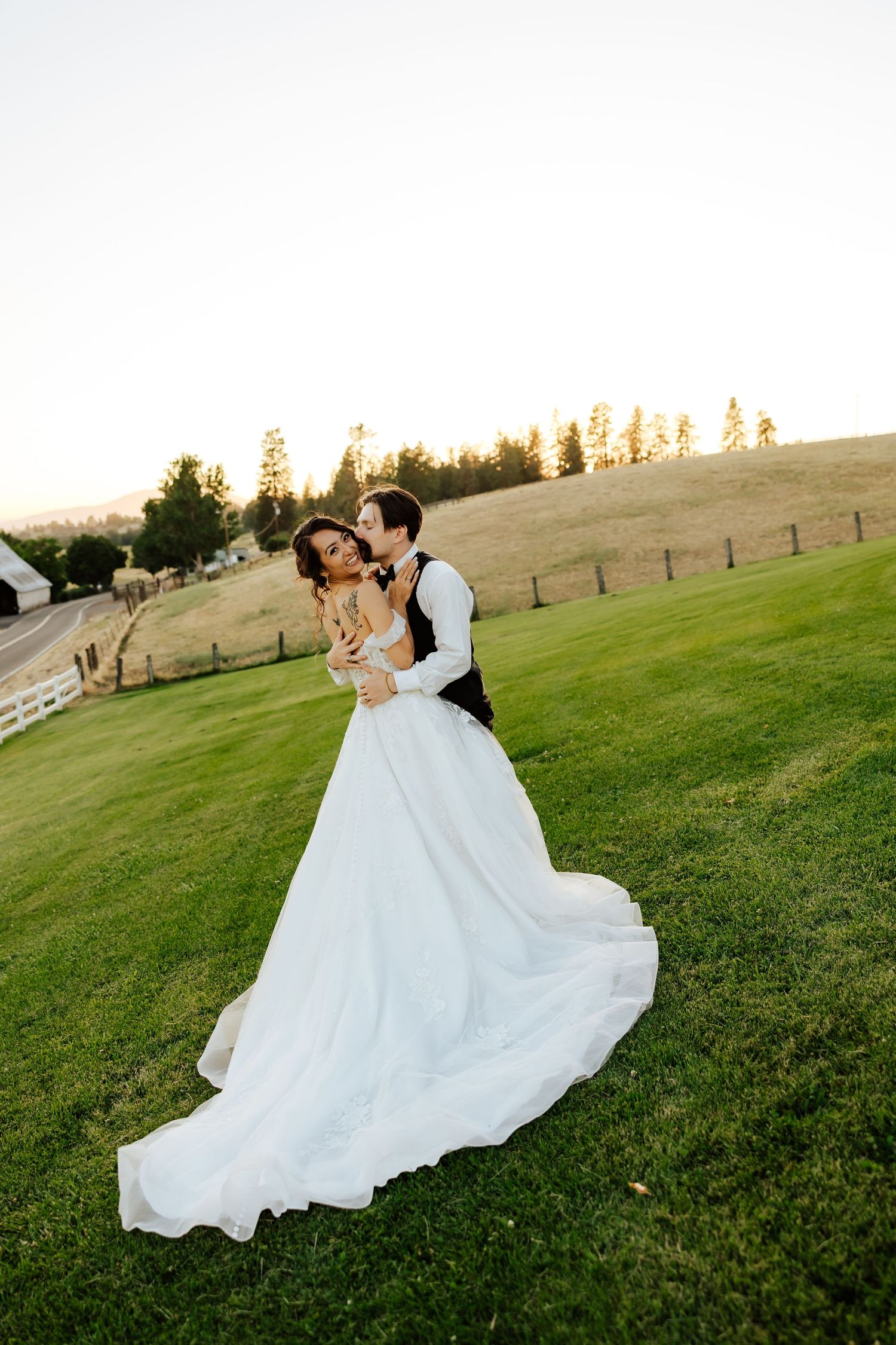 Groom embracing bride in flowing white gown on green hillside at sunset with trees in background.