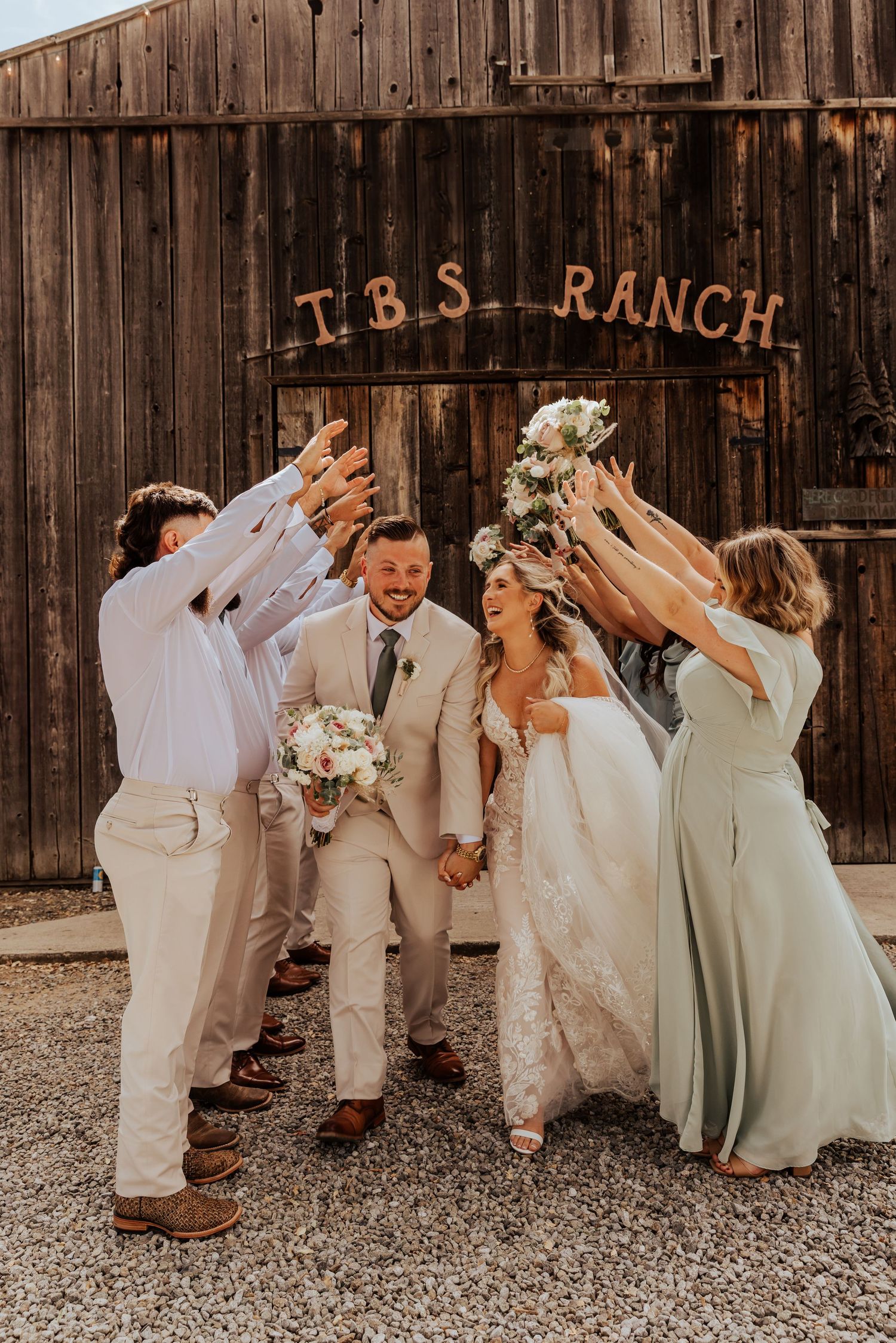 Wedding party posing playfully in front of rustic ranch barn wall.