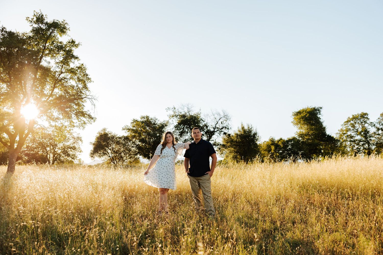 Couple walking hand in hand through tall golden grass field at sunset with trees in background.