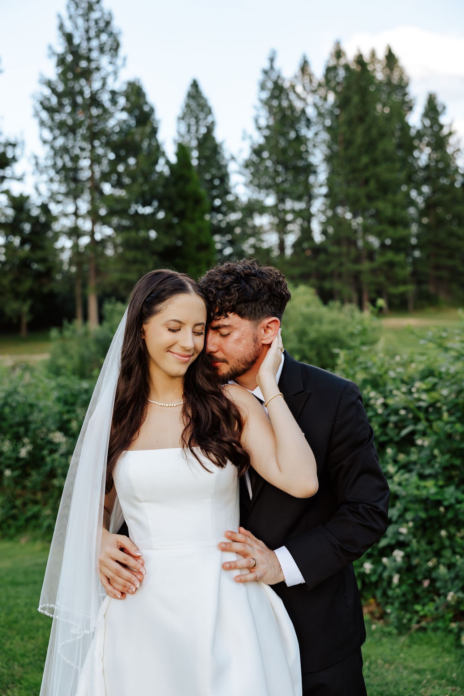 A groom in a black suit tenderly nuzzles a bride in a white strapless gown and veil outdoors.