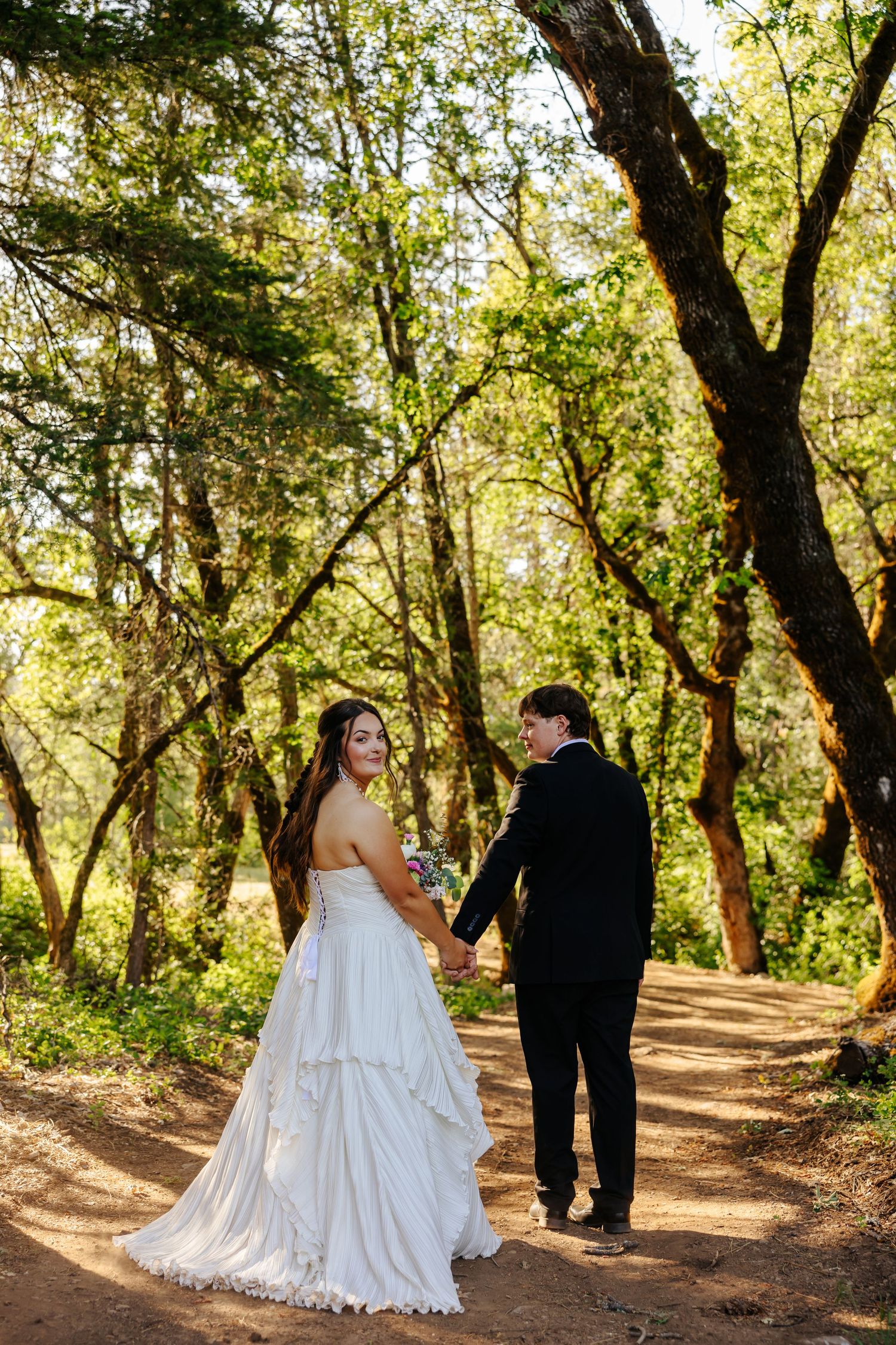 Bride and groom walk hand in hand along a sunlit forest path.
