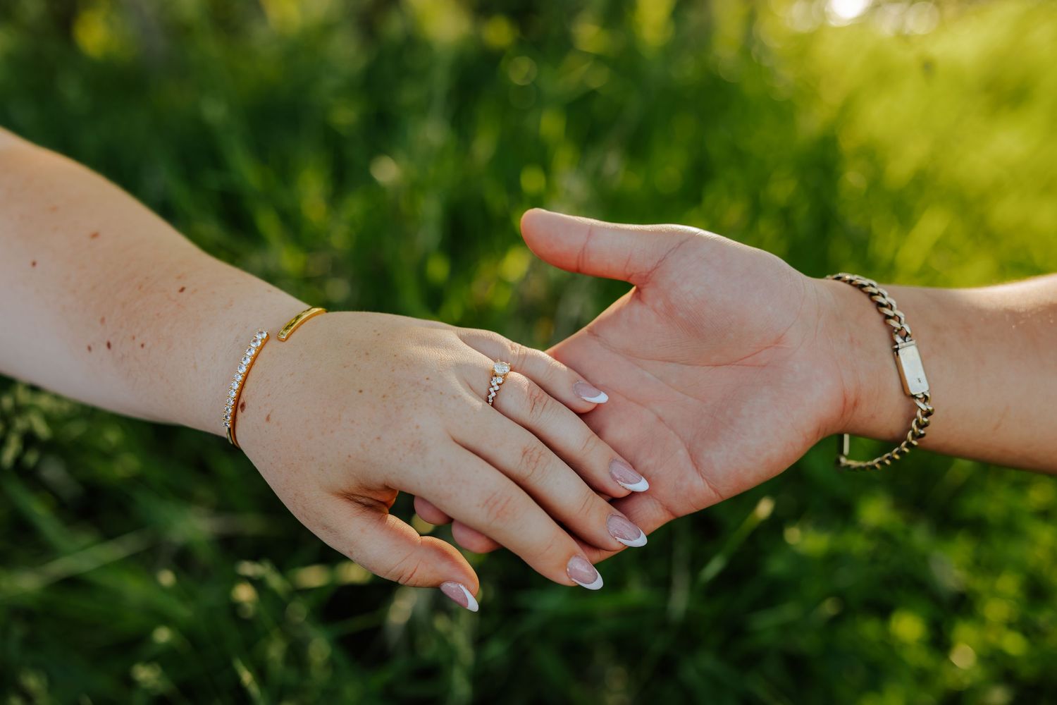 Two hands touching outdoors, adorned with gold bracelets and a ring, set against a lush green background.