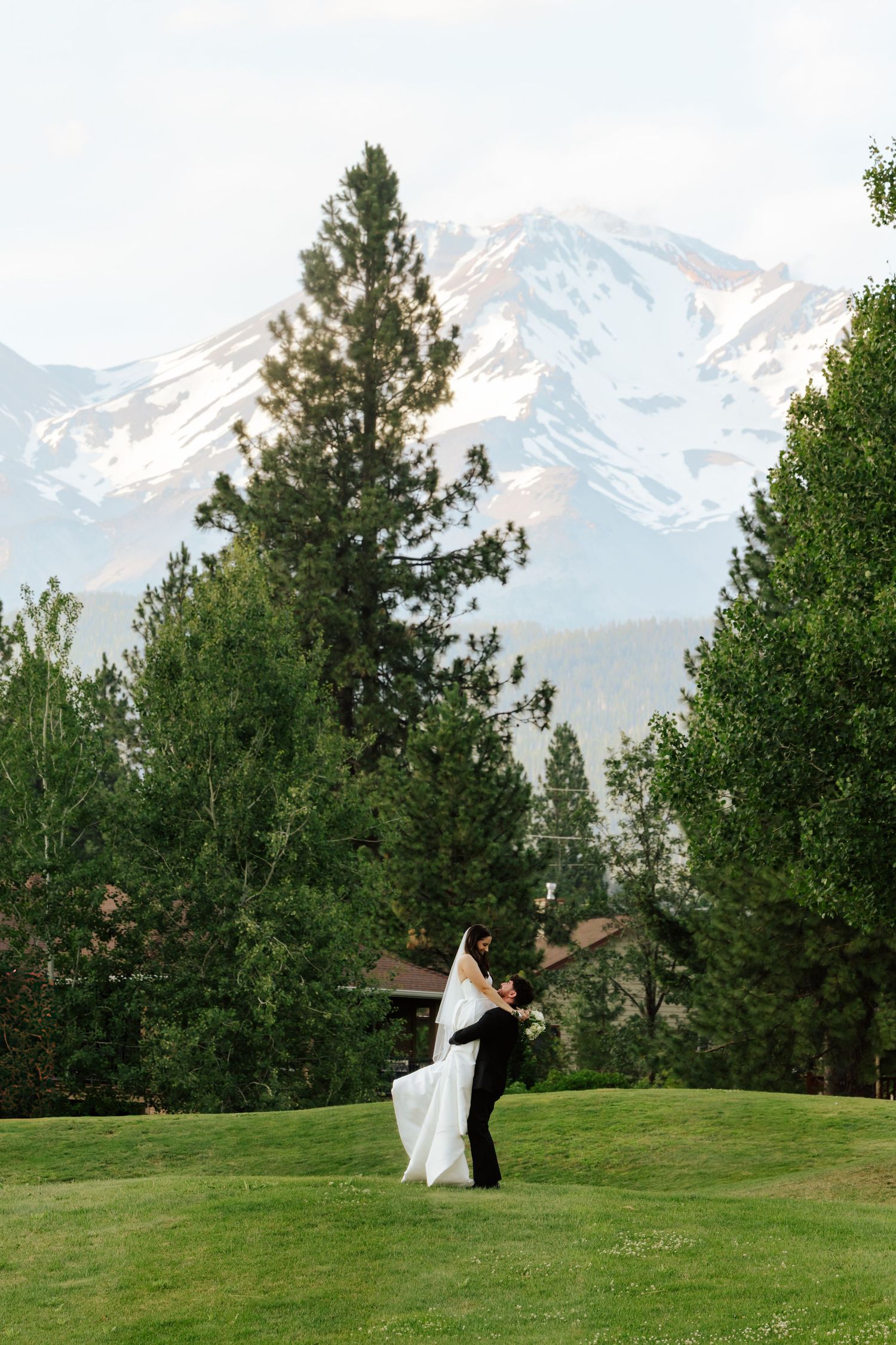 A bride and groom share a romantic embrace on a lush green lawn with snow-capped mountains and tall pine trees behind them.
