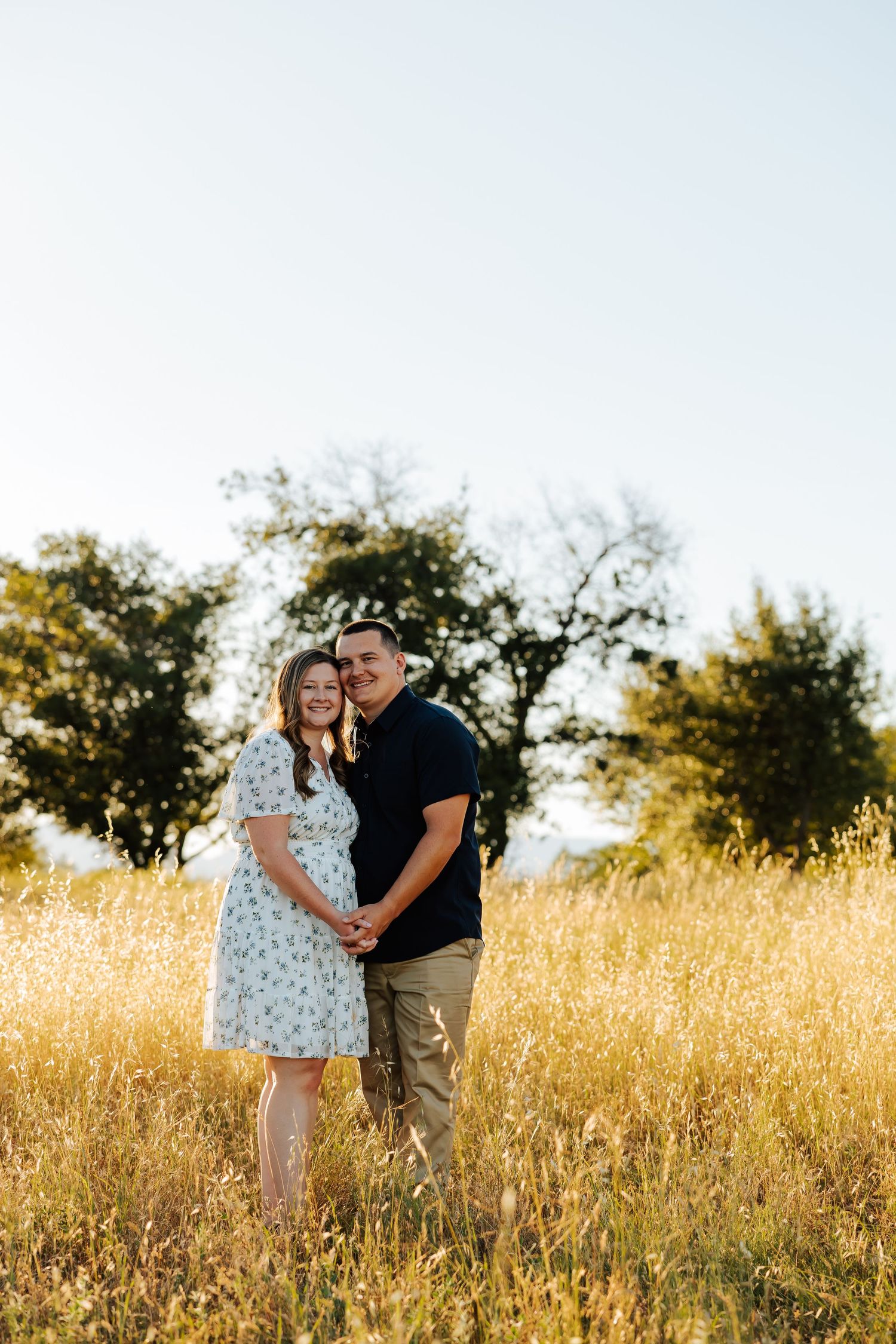 Couple smiling together in a golden sunlit field, woman in floral dress holding baby bump, trees in background.