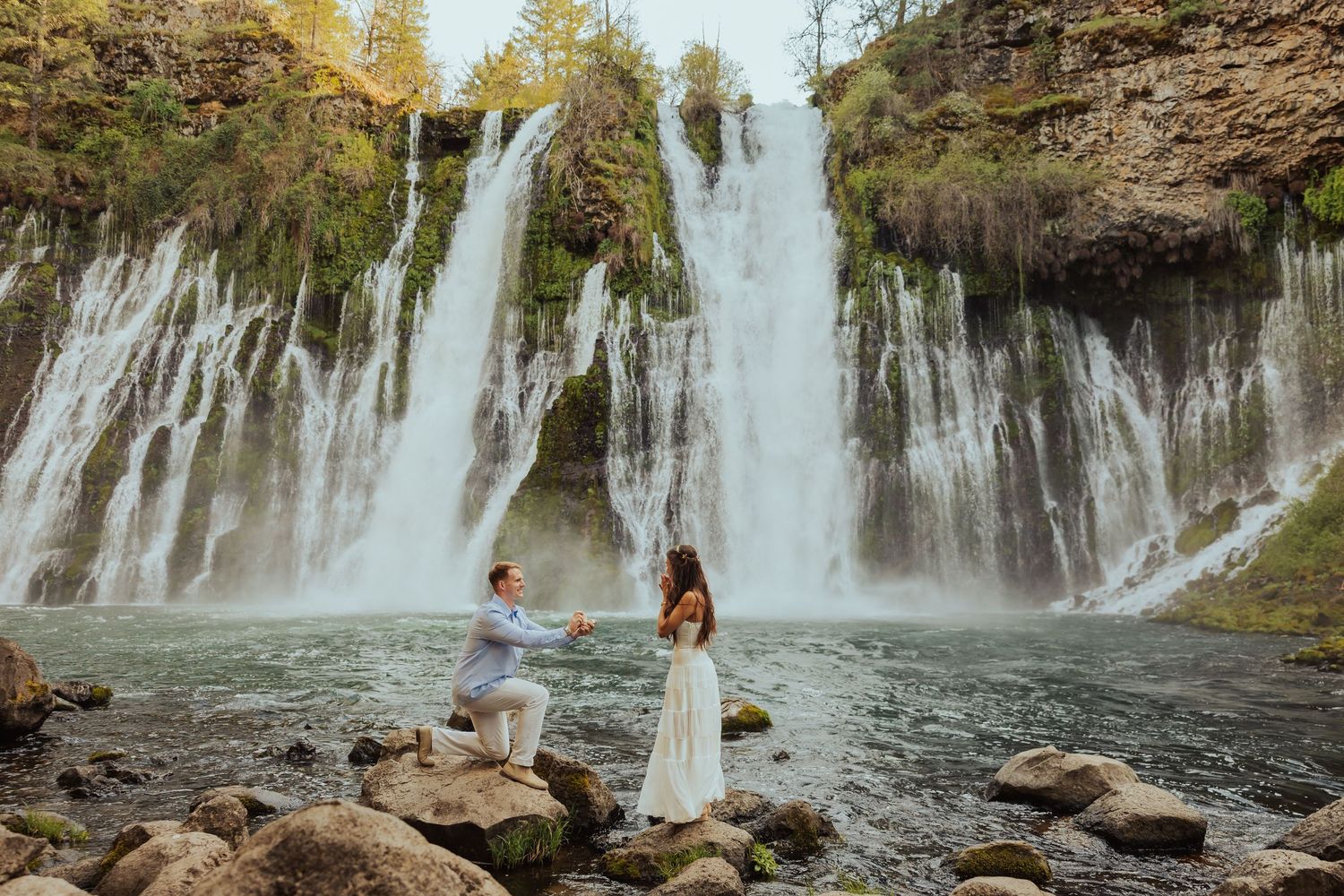 Man proposes to woman on rocky riverbank before stunning Burney Falls waterfall surrounded by autumn trees.