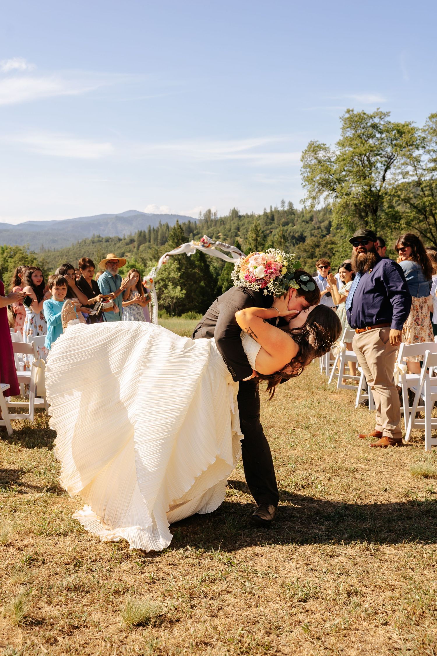 Groom dips bride for a romantic kiss at outdoor wedding ceremony with mountain views and guests cheering.
