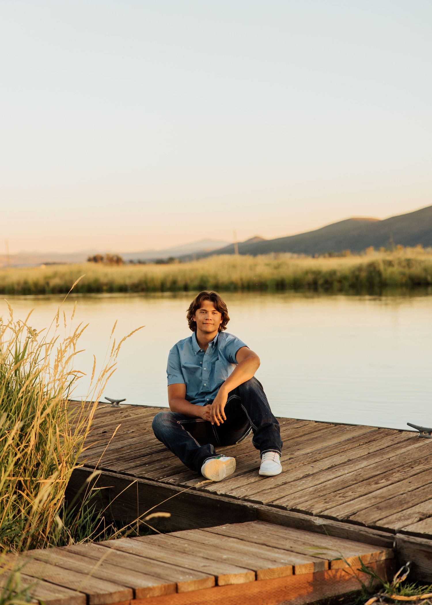 Person sitting on wooden dock by tranquil lake during sunset with mountains in background.